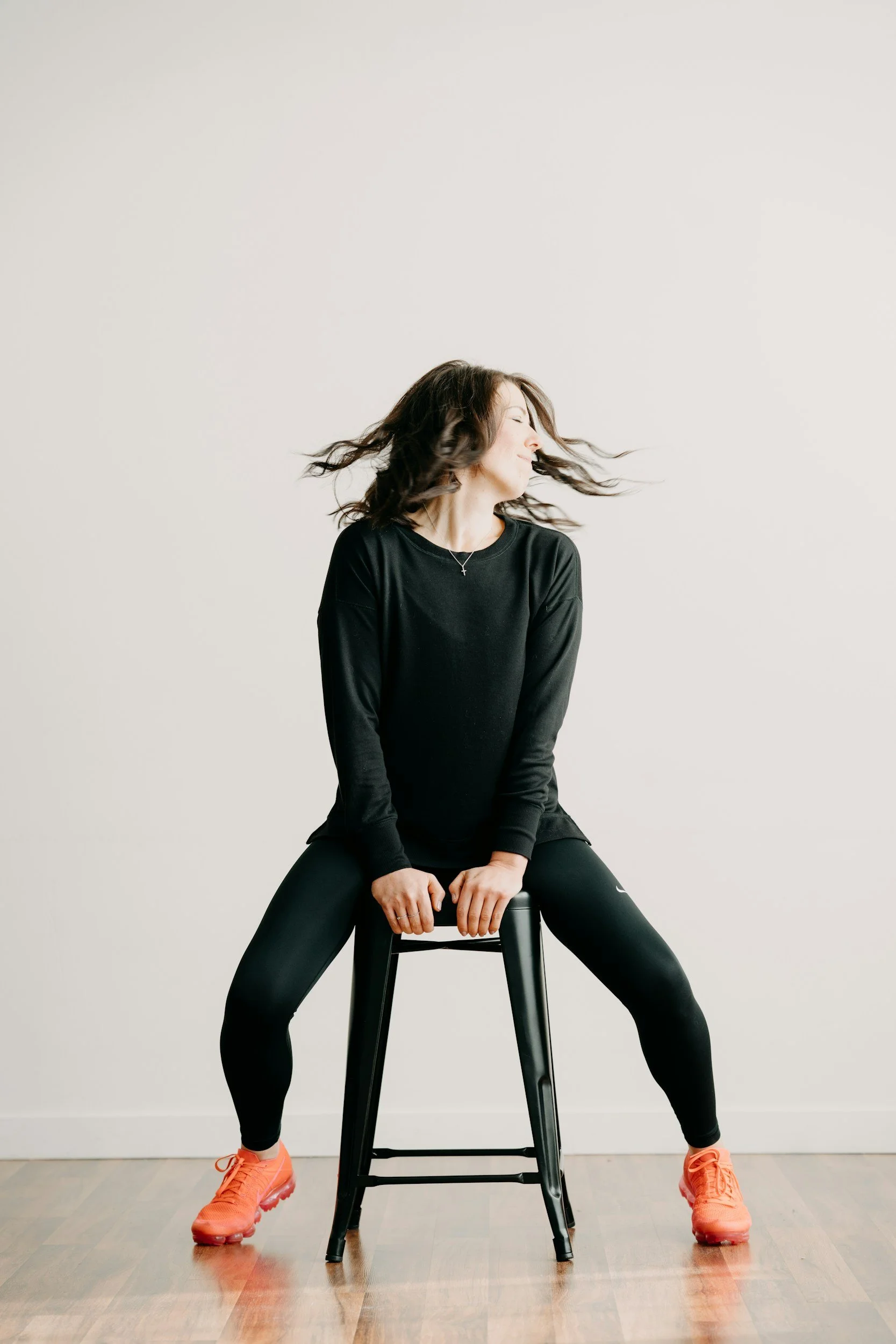 A woman wearing a black long-sleeve shirt, black sports leggings, and orange athletic shoes sitting on a black stool with her legs apart and hair blowing to the side against a plain white background.