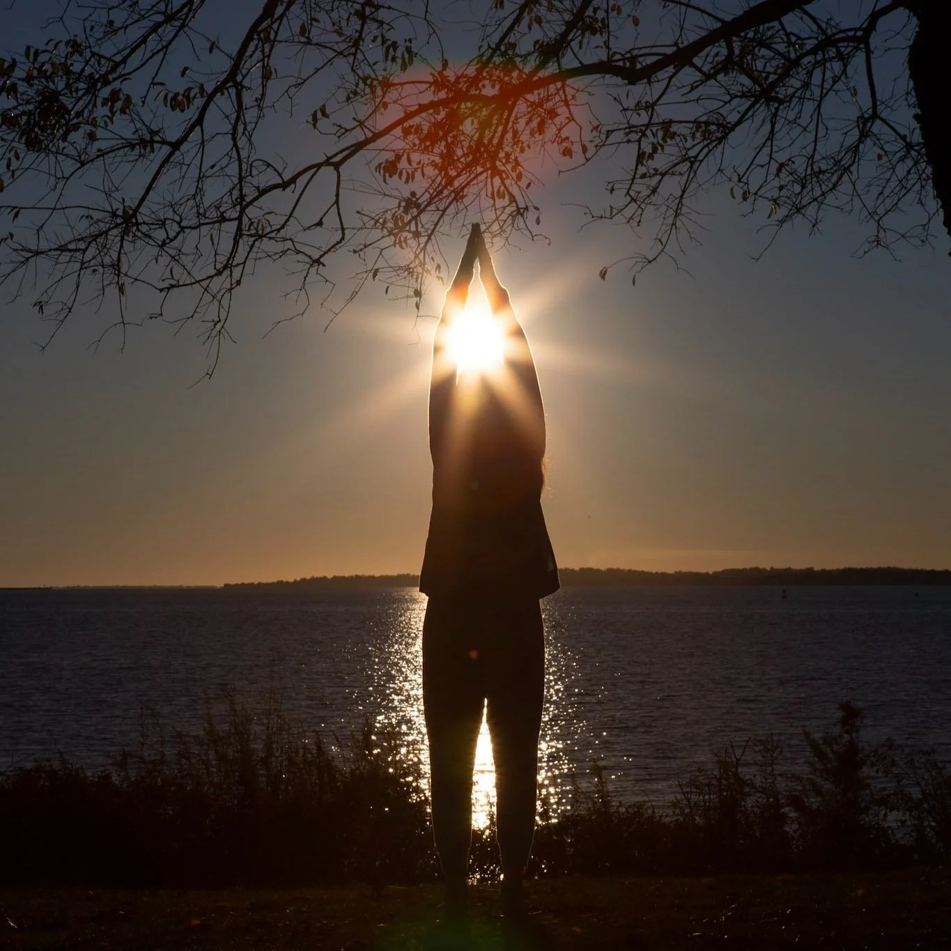 Silhouette of a person standing with hands pressed together above their head, creating a prayer pose, with the sun shining through their hands at sunset over a body of water.