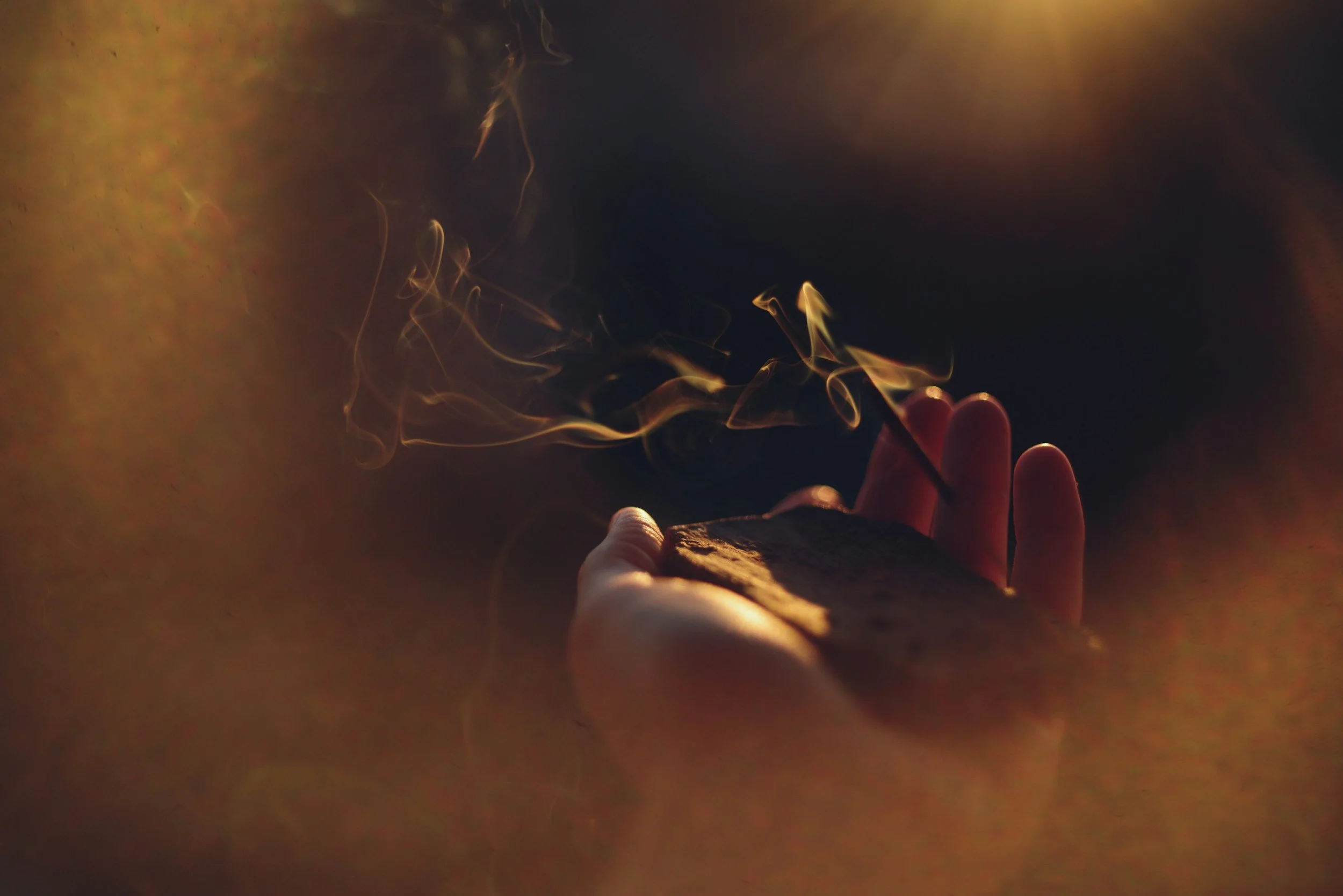 A close-up of a hand holding a piece of wood or similar object while delicately holding a lit incense stick, with smoke swirling upward against a dark, warm background with orange and brown hues.