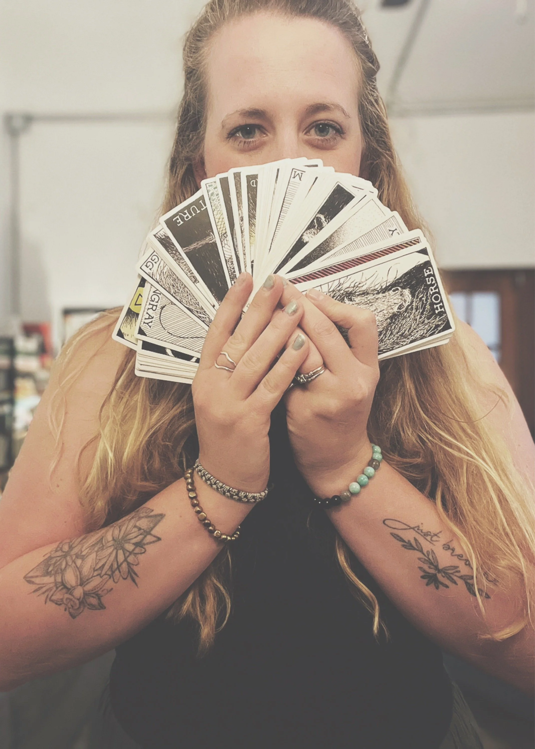 A woman with long, wavy blonde hair holding a fan of playing cards partially covering her face. She has tattoos on her arms and wears multiple bracelets. The background appears to be an indoor setting.