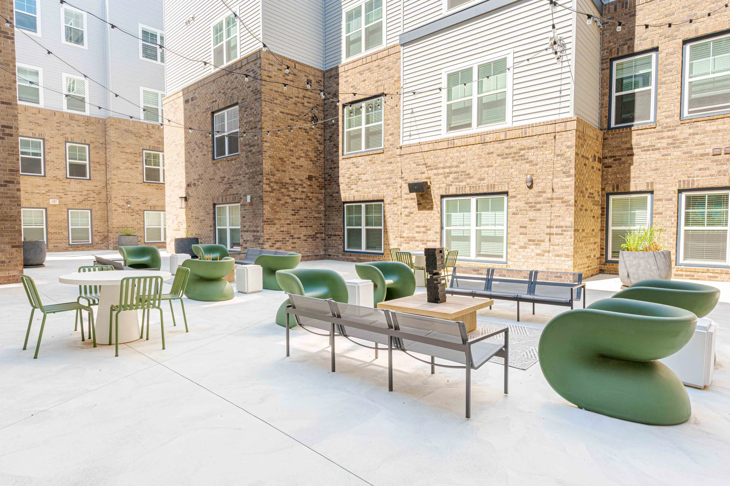 An outdoor patio area with modern green chairs, benches, and tables, surrounded by brick and white siding apartment buildings with string lights overhead.