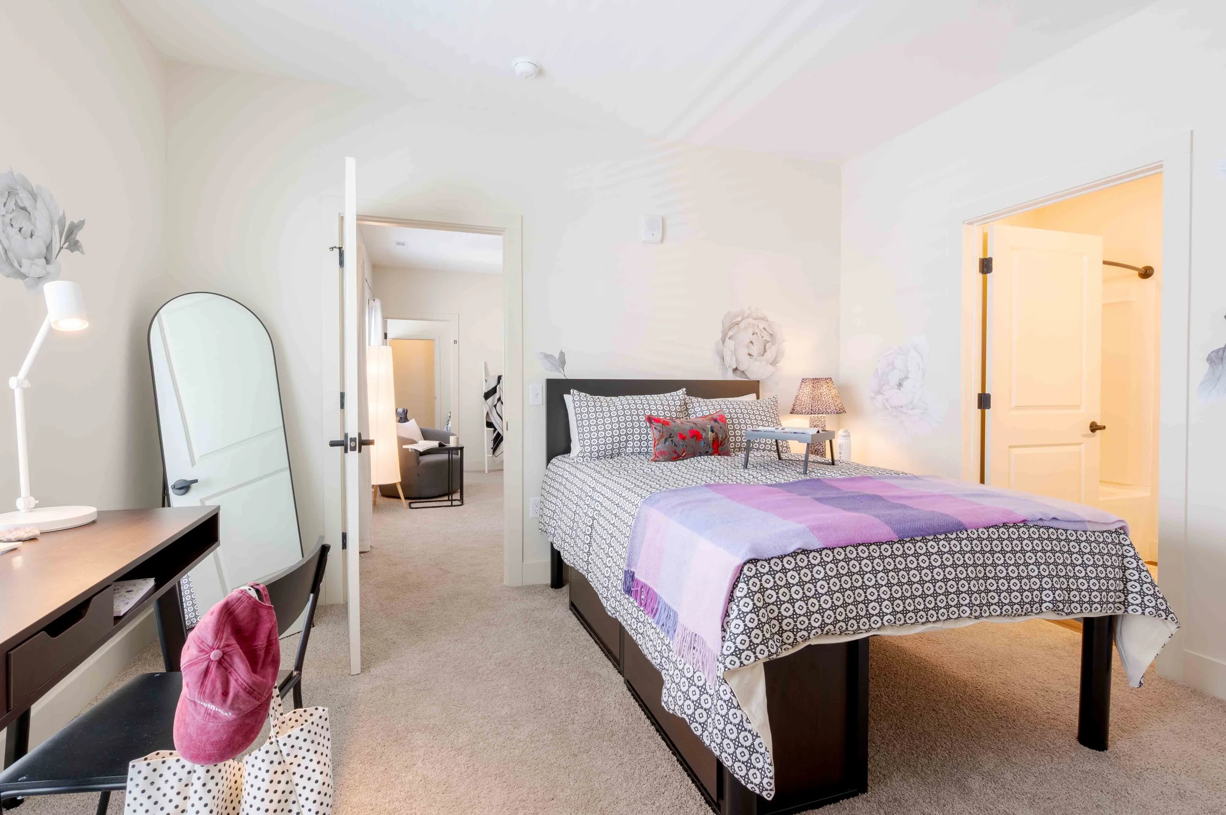 Interior of a bedroom with a bed, lamps, mirror, desk, and chairs, with an open door leading to another room.