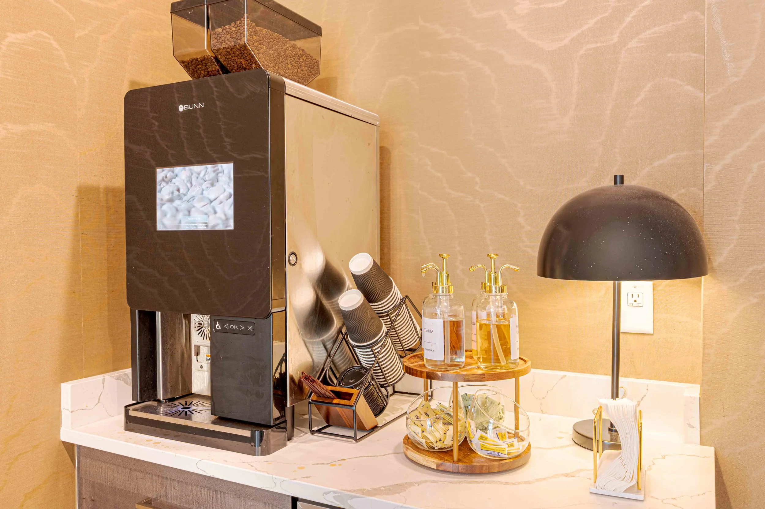 Coffee station with a BUNN coffee machine, disposable cups, a wire basket with tea bags, bottles of vanilla and caramel syrup, a black lamp, and a beige wall in the background.
