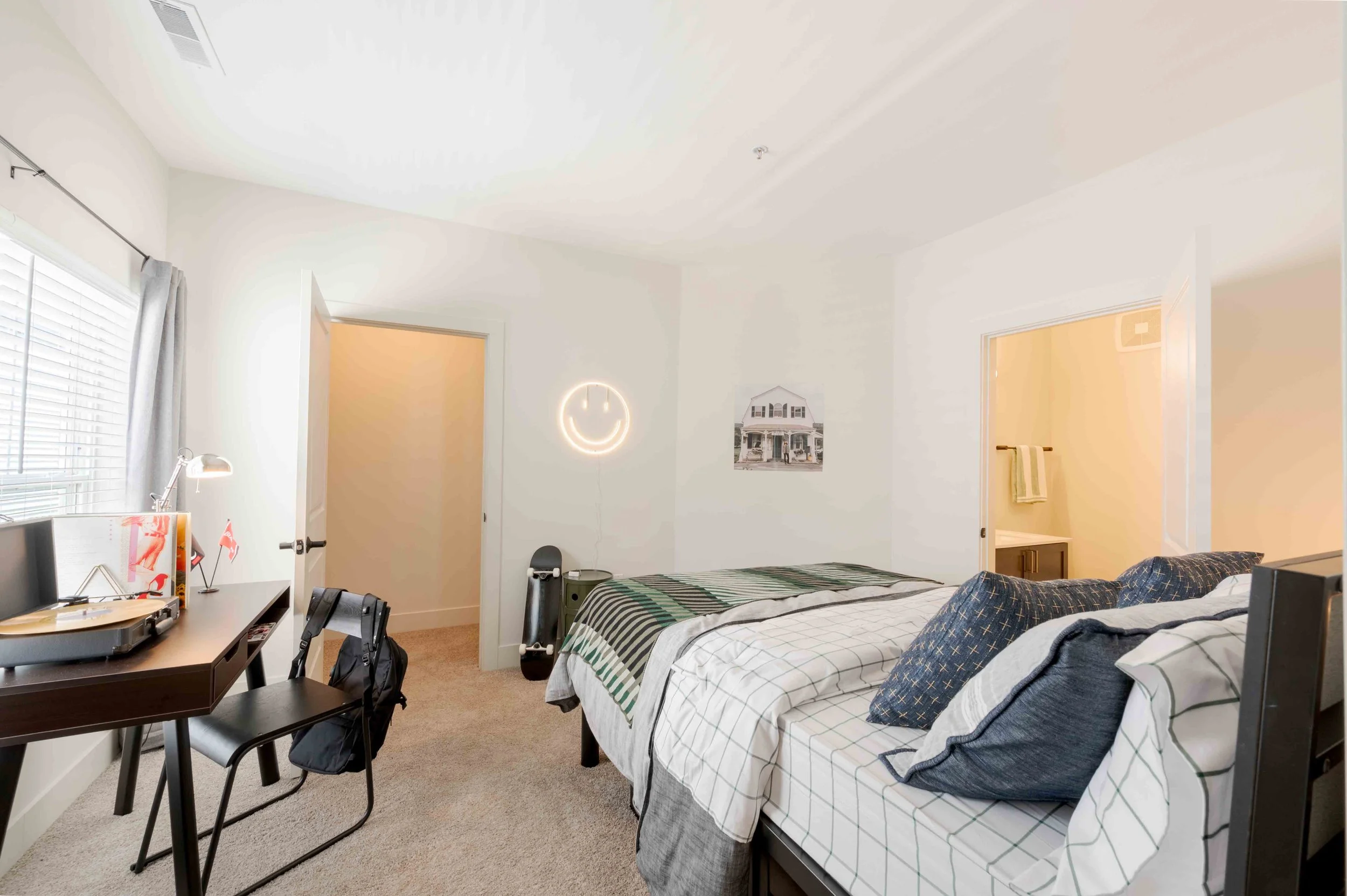 A bedroom with two beds, each with checkered bedspreads, pillows, a window with blinds and curtains, a neon smiley face wall light, a wall photo of a house, a desk with a lamp and gadgets, and some chairs and bags.