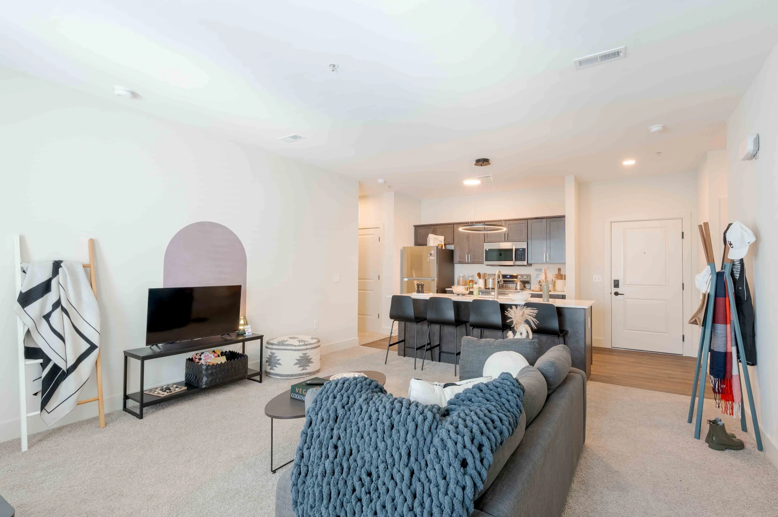 Living area and kitchen in an apartment or home, with a TV on a black stand, a cozy gray couch with a chunky-knit blanket, a round coffee table, and various decorations. The kitchen has dark cabinets, white countertops, a refrigerator, microwave, and