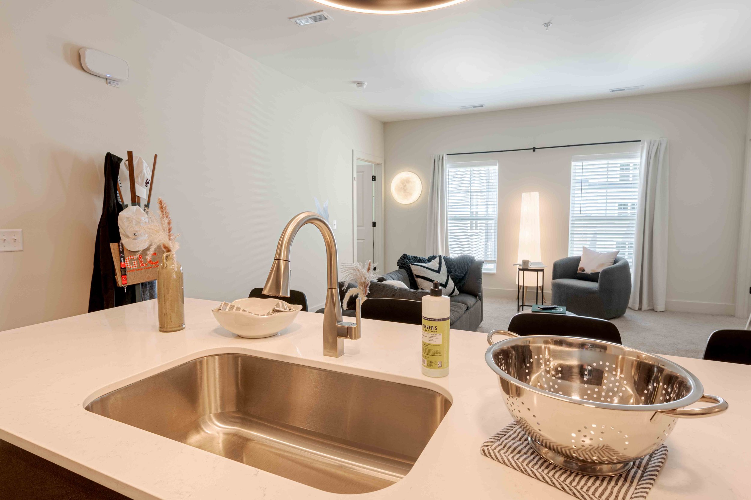 View of a modern living room and kitchen area. The kitchen countertop has a stainless steel sink with a brass faucet, a colander, and a bottle of soap. In the background, there are two gray chairs, a small table, and white curtains on the windows. Th