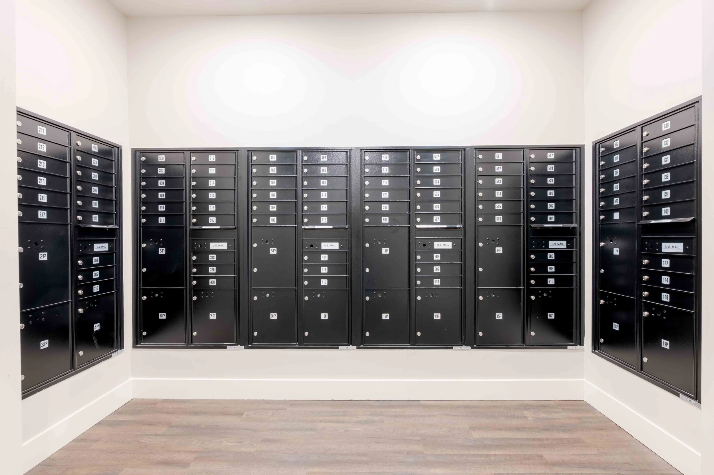 Bank of black mailboxes aligned in a room with white walls and wood flooring.
