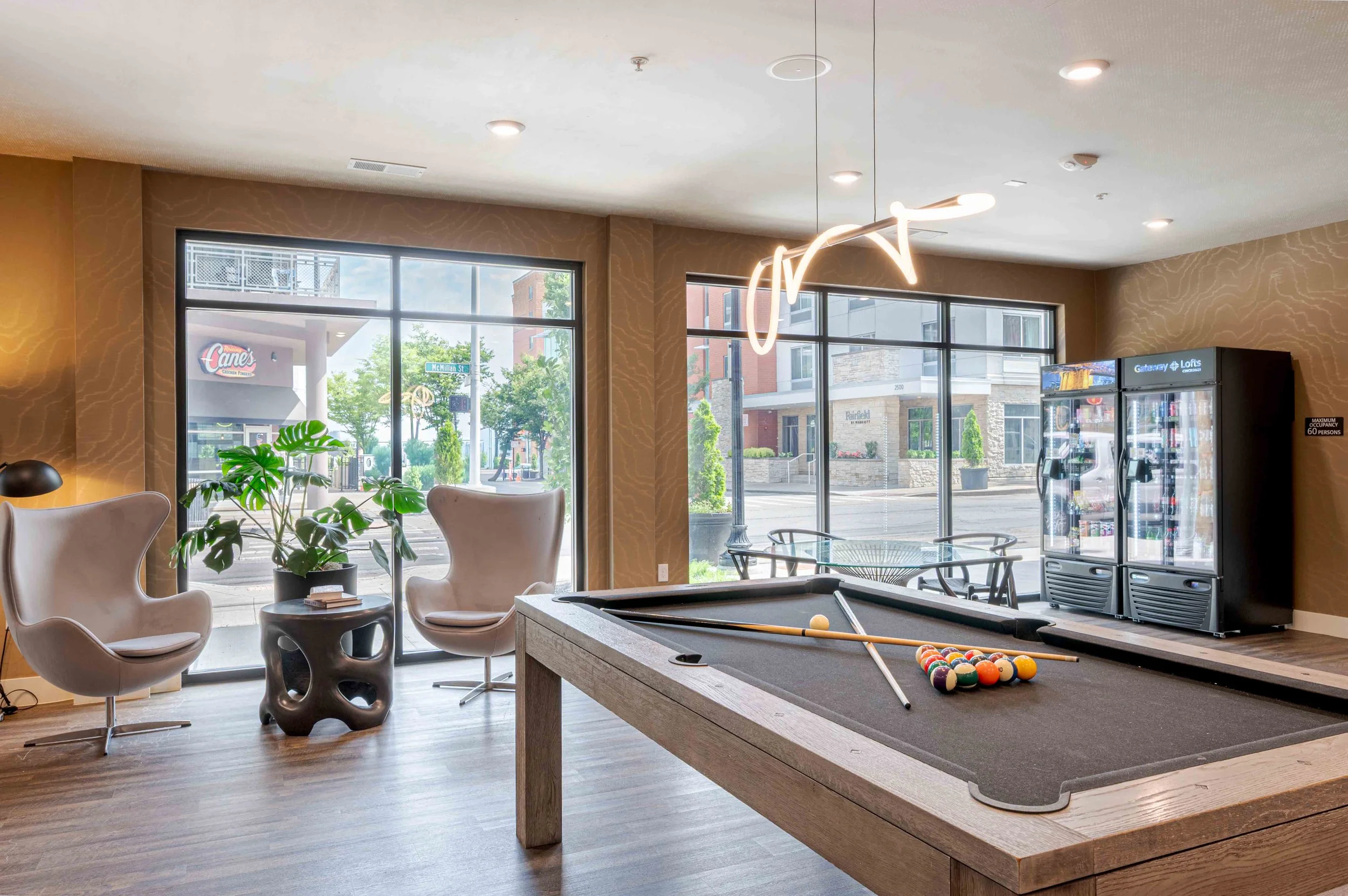 Interior of a modern lounge or common area with large windows, a pool table, comfortable white chairs, a plant, and a refrigerator with beverages, illuminated by contemporary hanging light fixtures.
