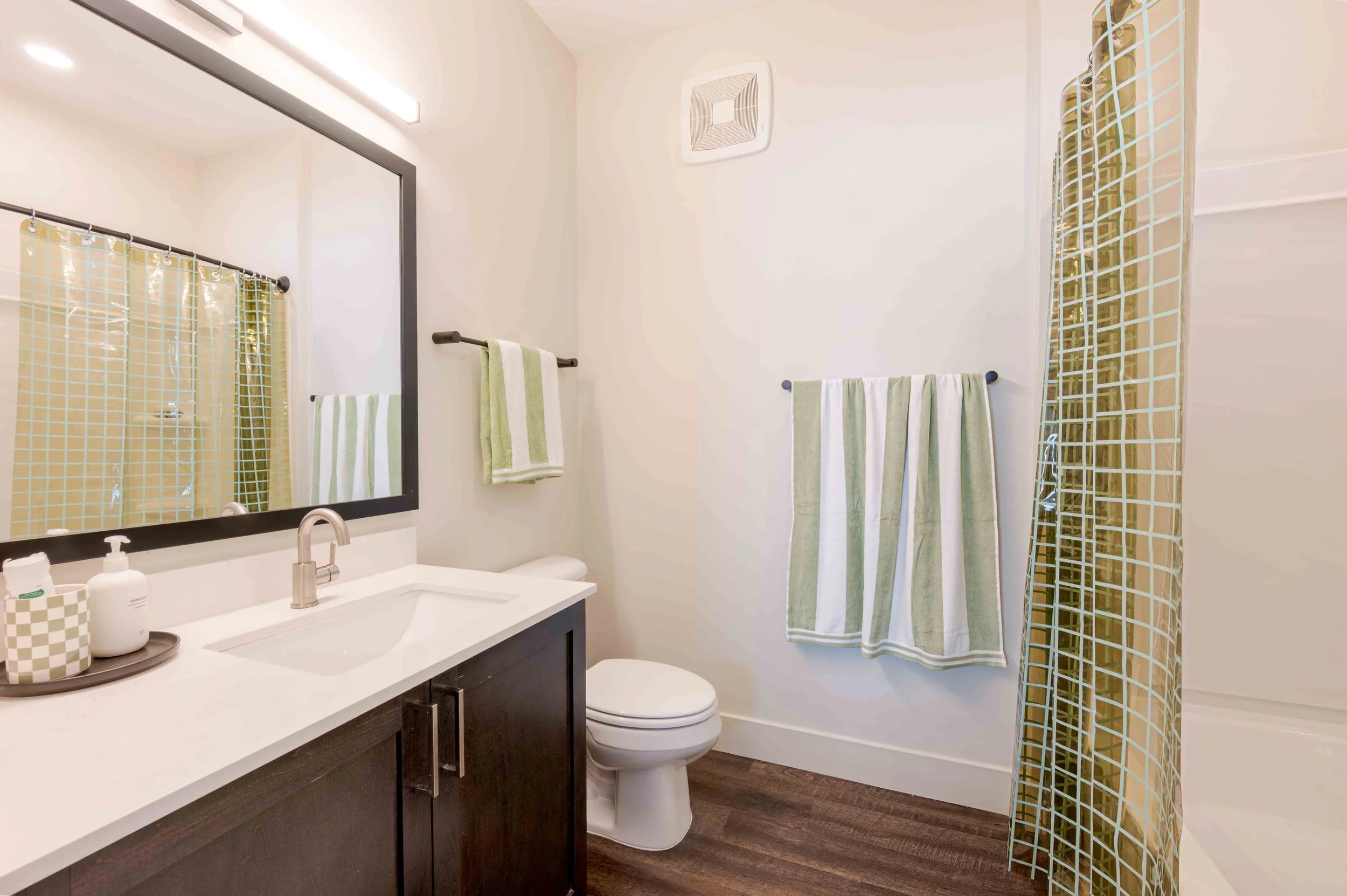 A bathroom with a dark cabinet vanity, a white countertop, and a large mirror with a gray frame. The mirror reflects a shower curtain with a yellow and green checkered pattern. There are light green and white towels on a black towel bar and a smaller