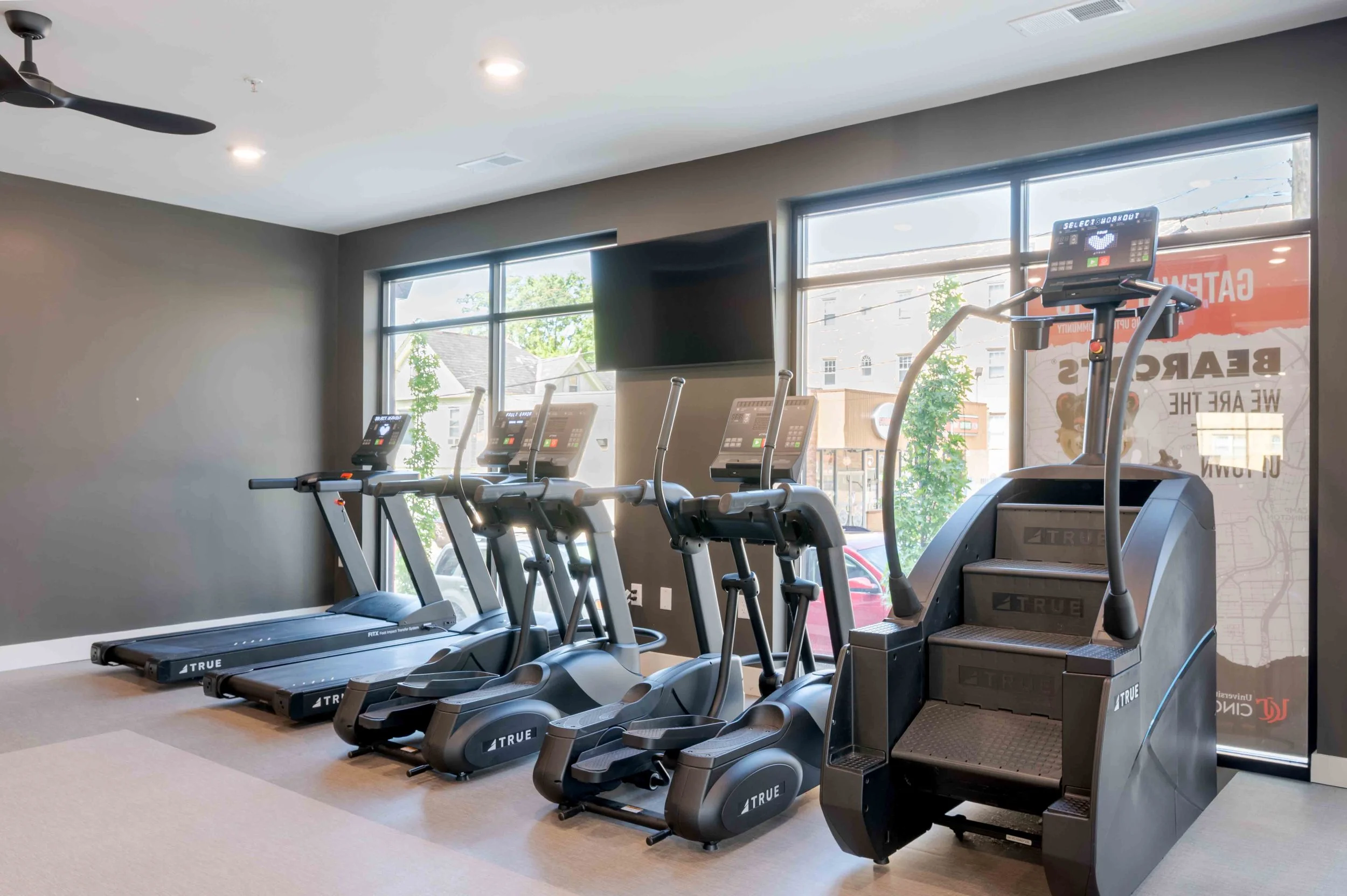 Empty gym with multiple treadmills facing large windows with a view of trees and buildings outside.