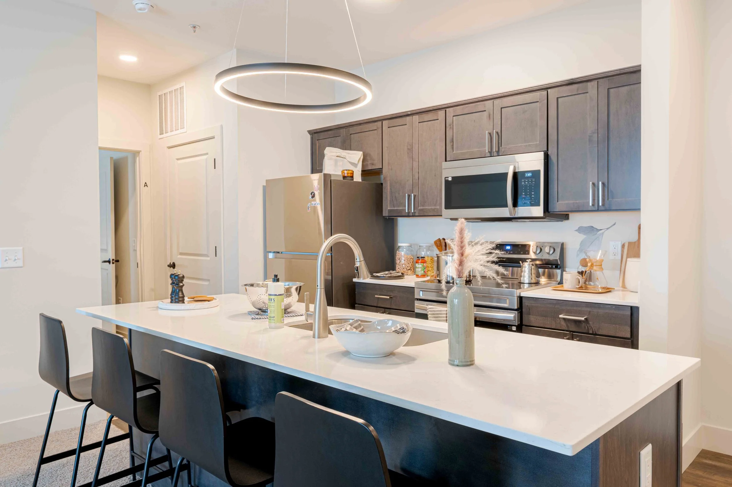 Modern kitchen with white island table, black chairs, overhead circular LED light, gray wooden cabinets, stainless steel microwave, oven, and fridge, decorative vases, and kitchen items on the counter.