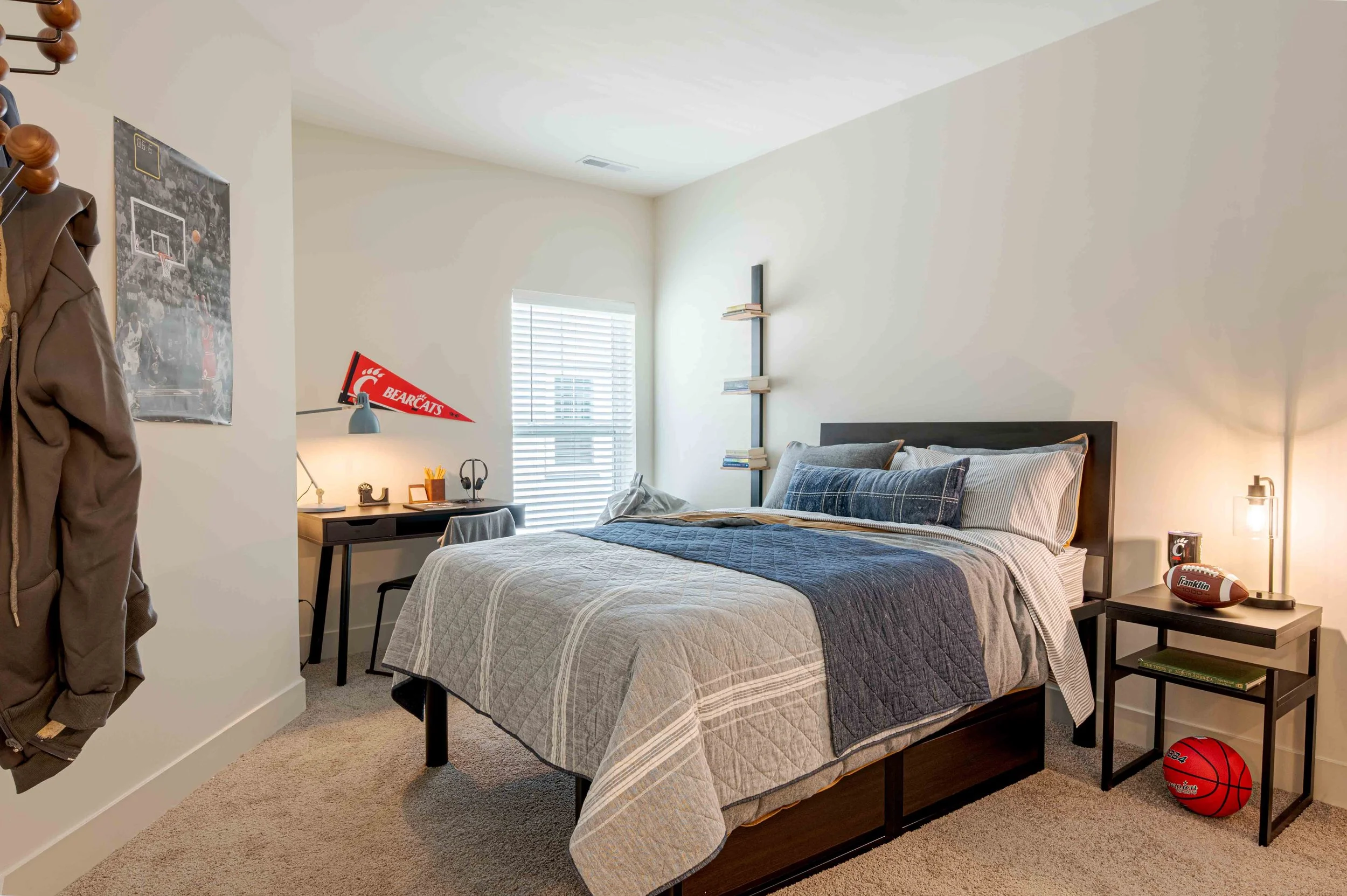 A bedroom with a bed, nightstand, and window with blinds. The bed has pillows and a quilted bedspread. A shelf with books is mounted on the wall, and a basketball is on the floor near the nightstand. A sports pennant that says 'BEARCATS' and a flag a
