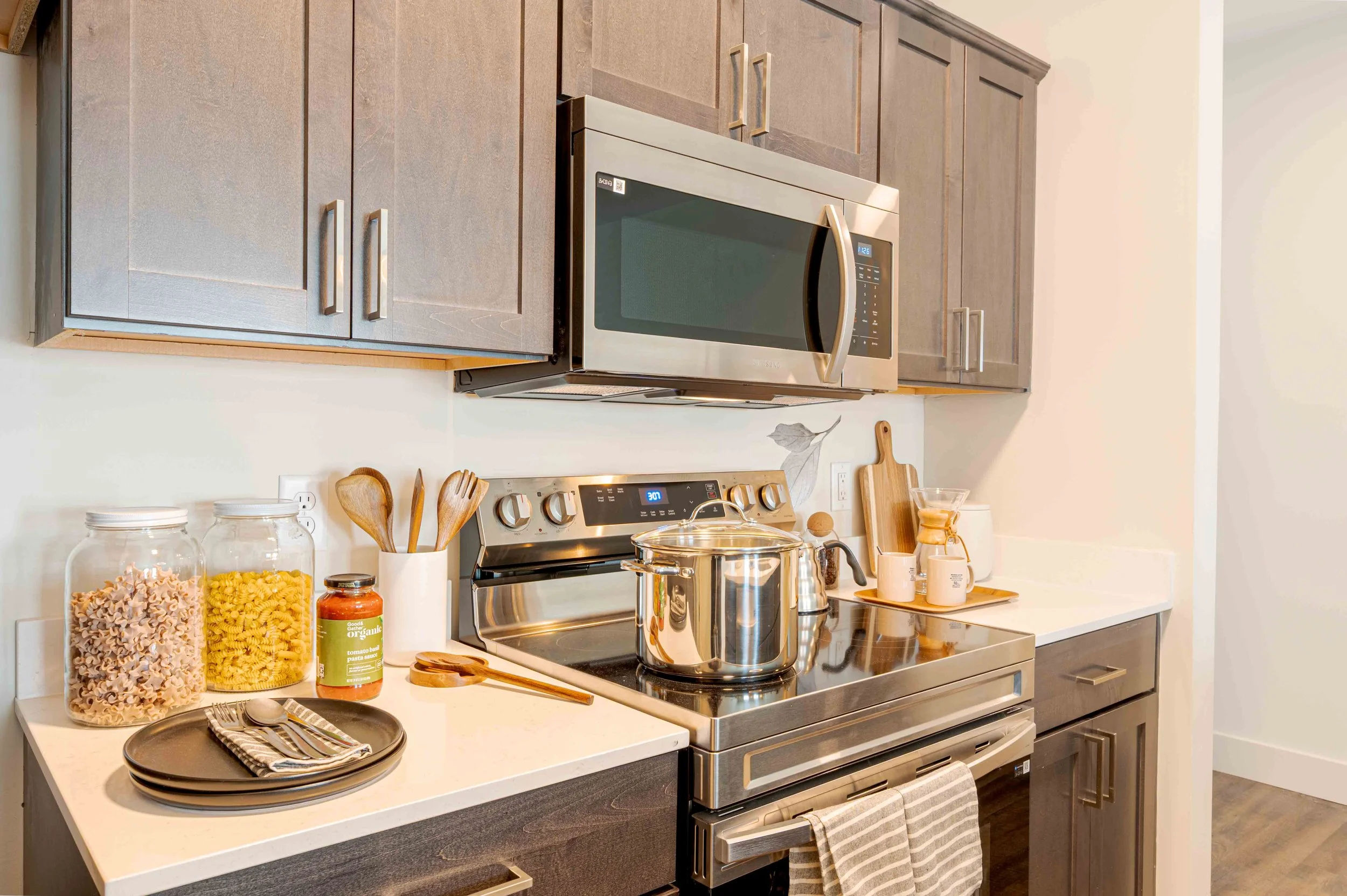 Modern kitchen with dark wood cabinets, stainless steel microwave and stove, glass jars with pasta, a pot on the stove, and various utensils and containers on the countertop.