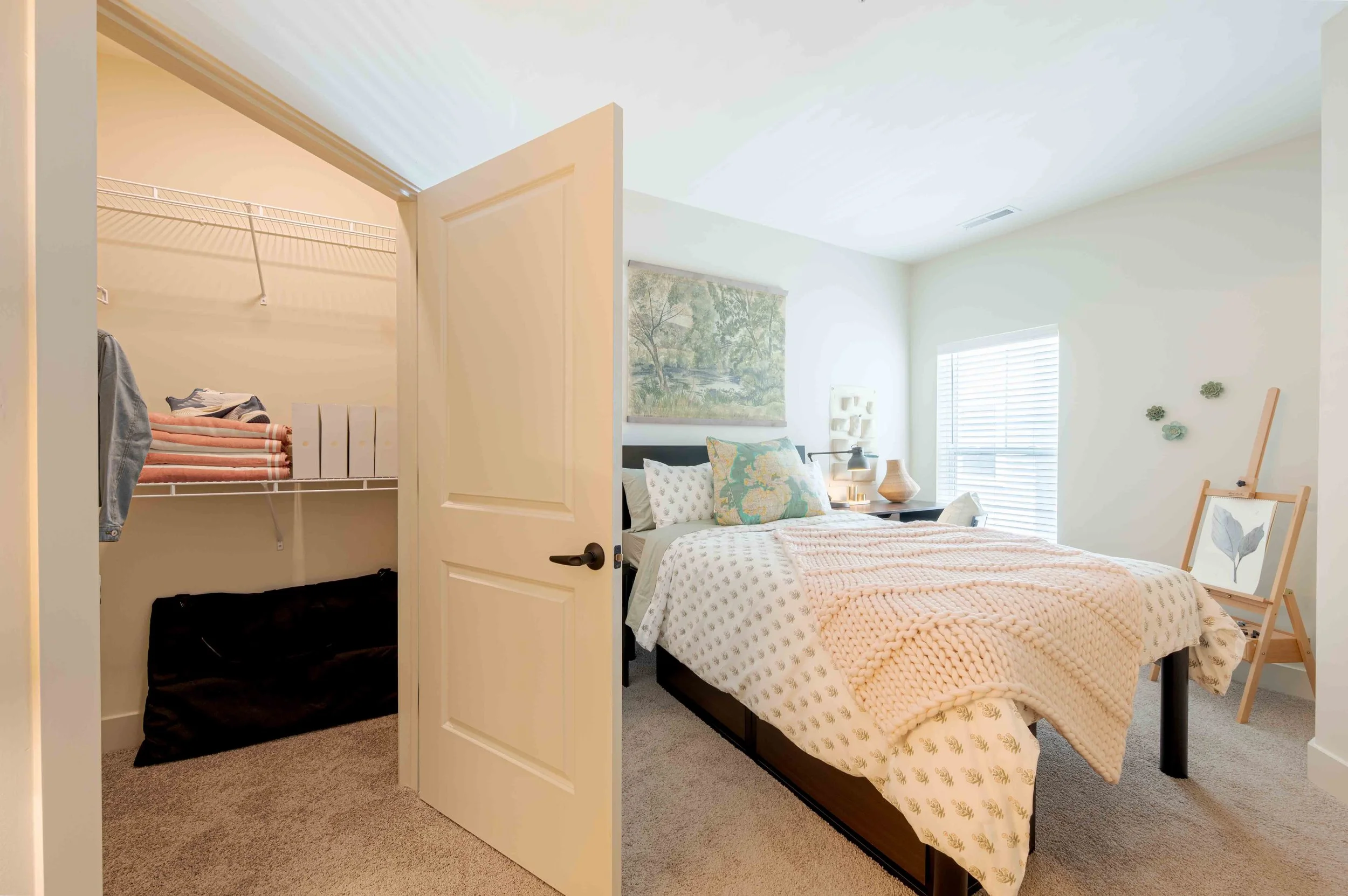 A bedroom with a bed, nightstand, lamp, and artwork on the wall, with a closet visible through an open door.