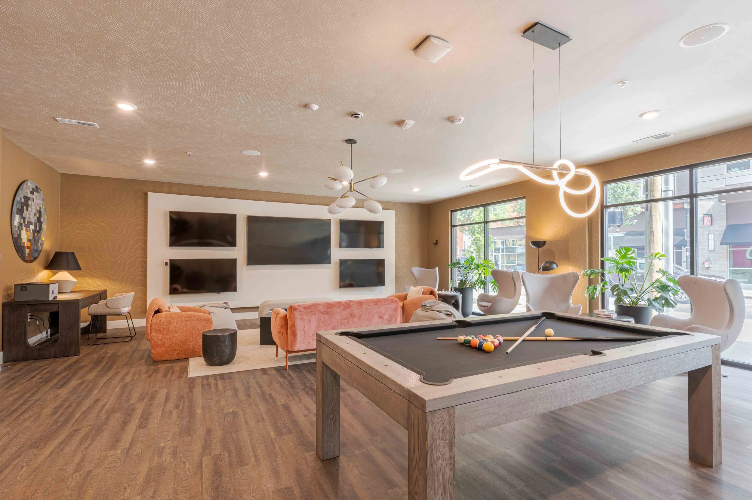 A modern entertainment room featuring a pool table in the foreground, a wall-mounted flat-screen TV with multiple screens, a contemporary chandelier, comfortable chairs, plants, and large windows letting in natural light.