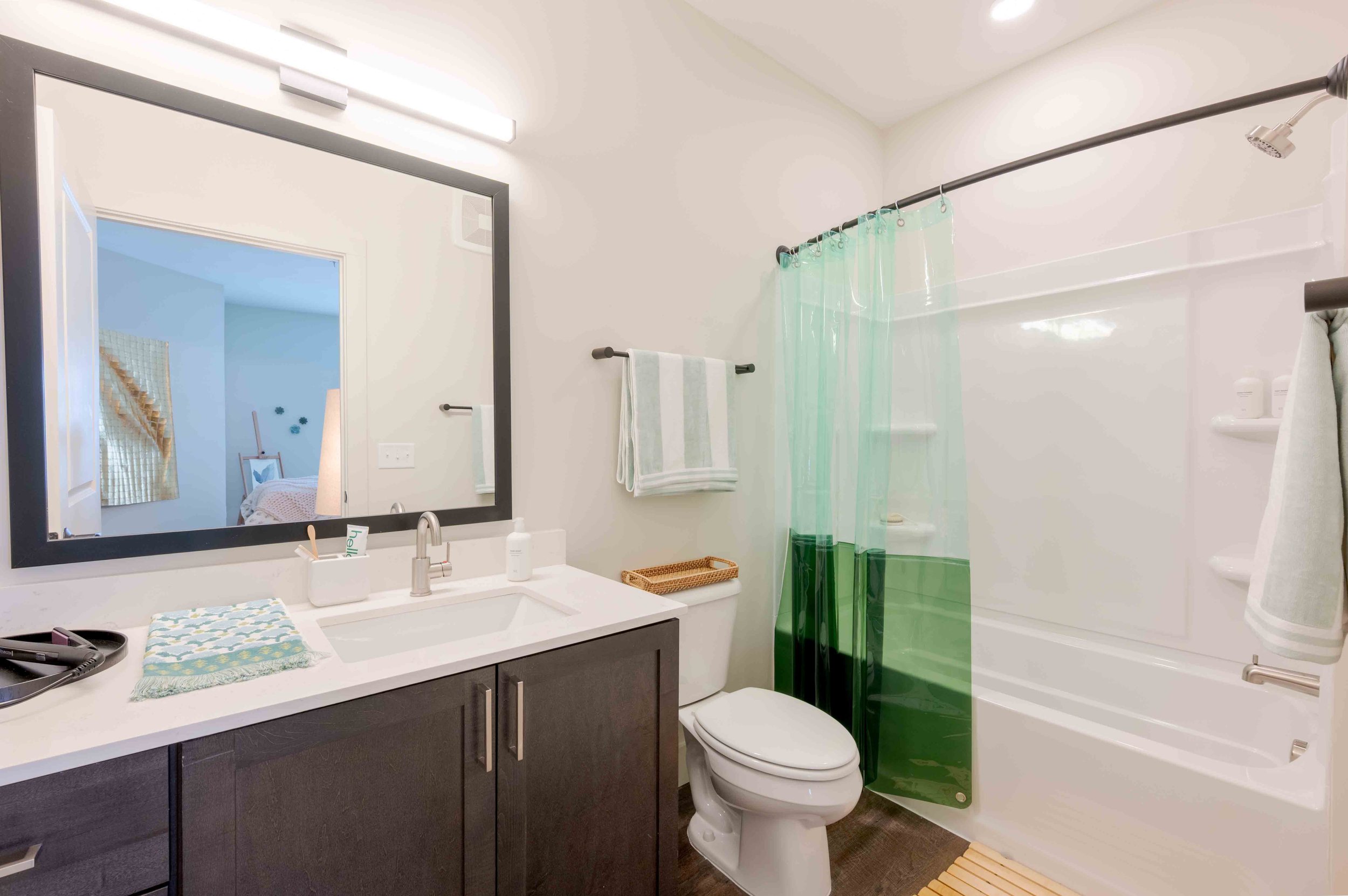 A bathroom with a vanity mirror, a sink with soap and toothbrush holder, a dark cabinet, a toilet, a shower with a green shower curtain, and white walls.
