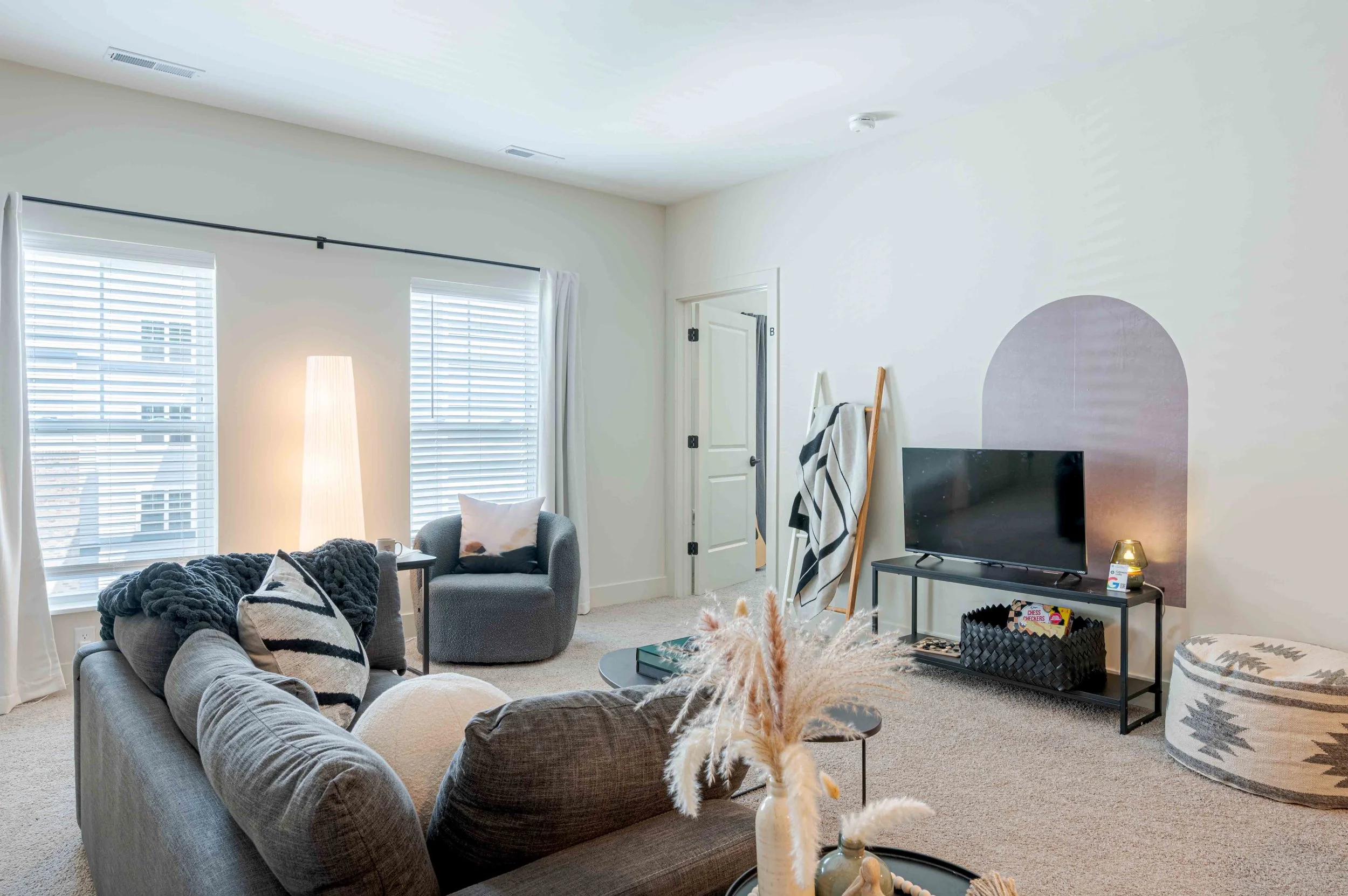 Living room with gray couch, armchair, black TV, coffee table, and window with blinds, decorated in neutral tones.