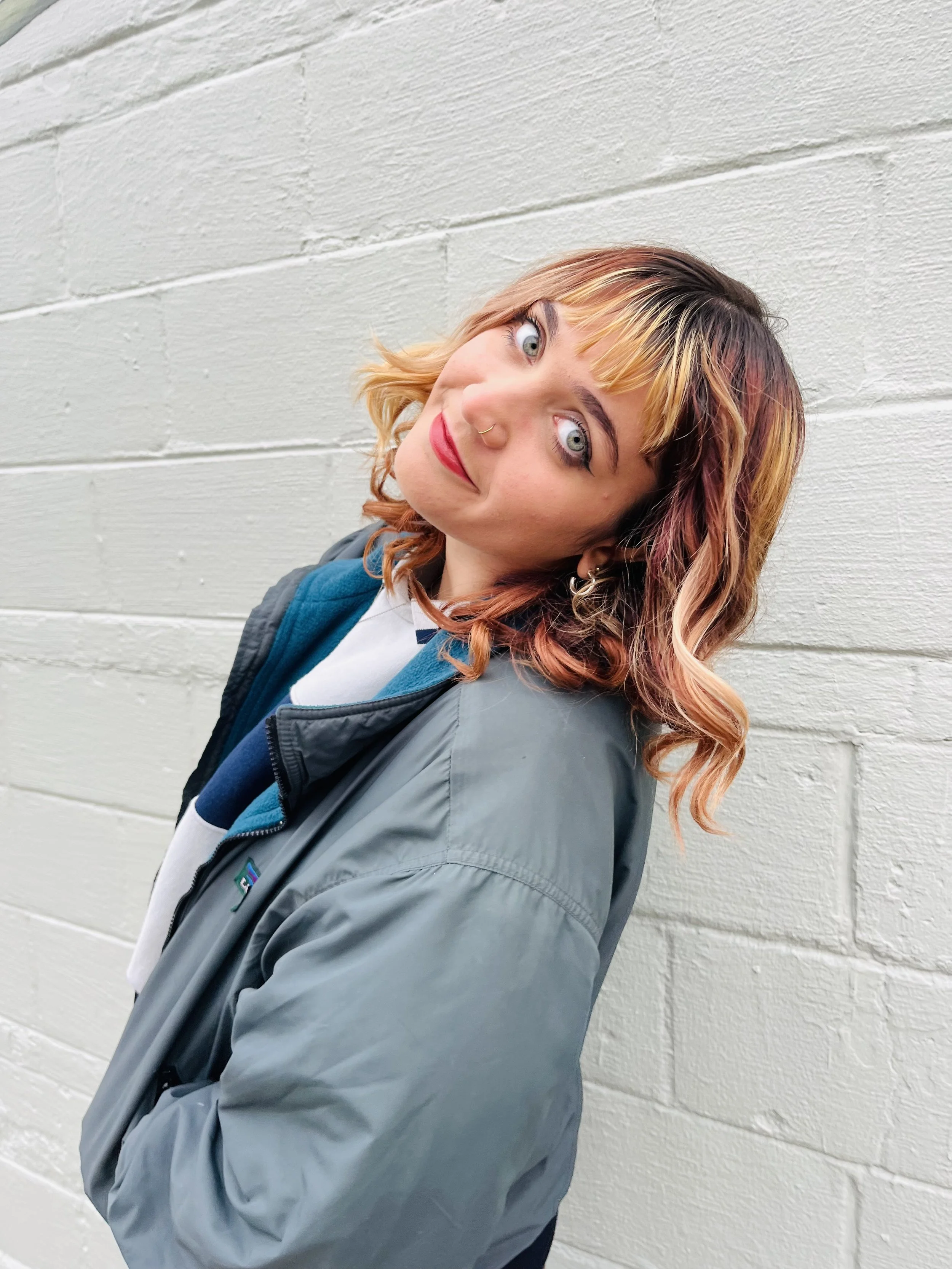 A young woman with wavy, reddish-blonde hair with darker roots, wearing a nose ring and striking red lipstick, leaning against a white brick wall, dressed in a light gray Patagonia jacket over a white shirt with blue accents.