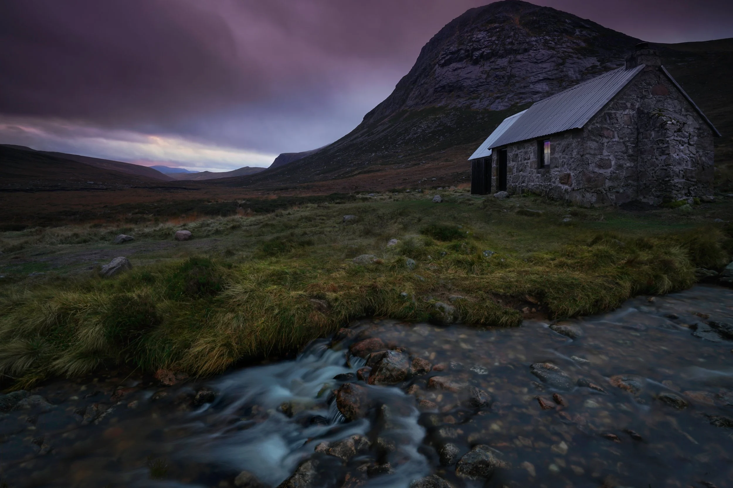 A small stone cabin with a metal roof nestled in a mountain landscape with a small stream in the foreground and dark, cloudy sky above