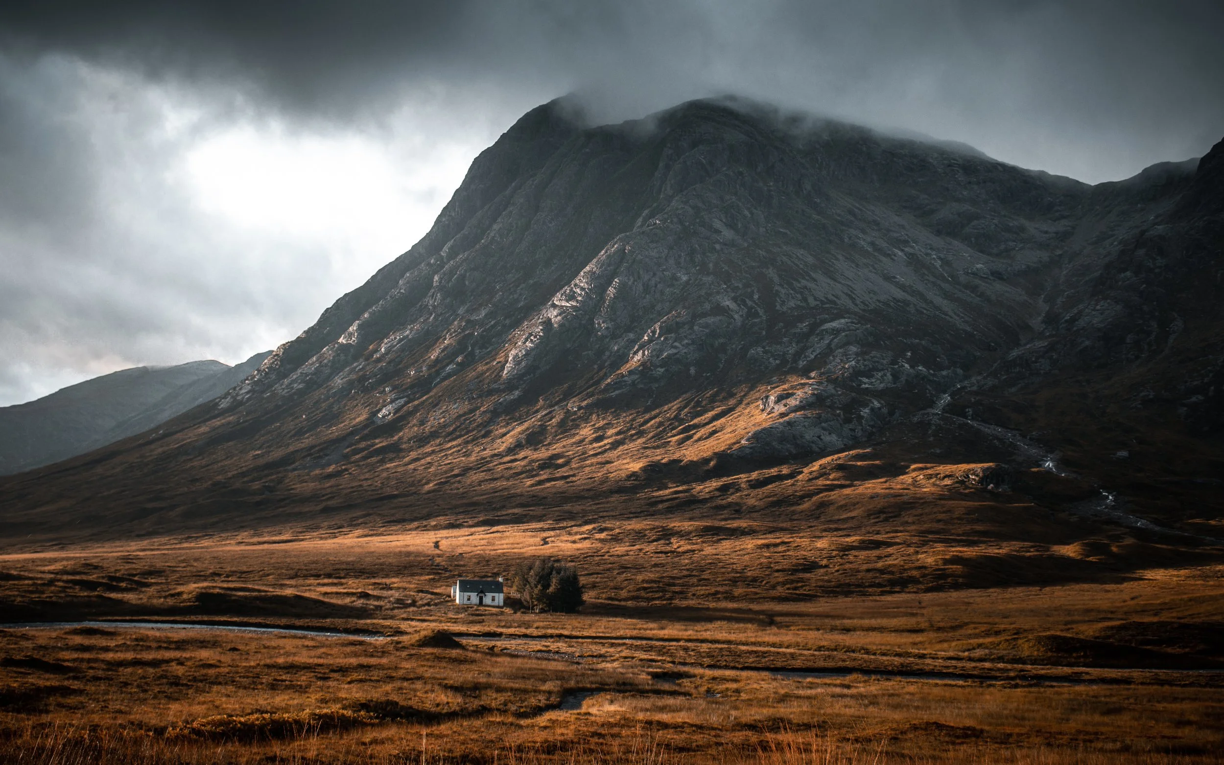 A small white house in a vast, open landscape with a large, rugged mountain in the background, overcast sky with dark clouds.