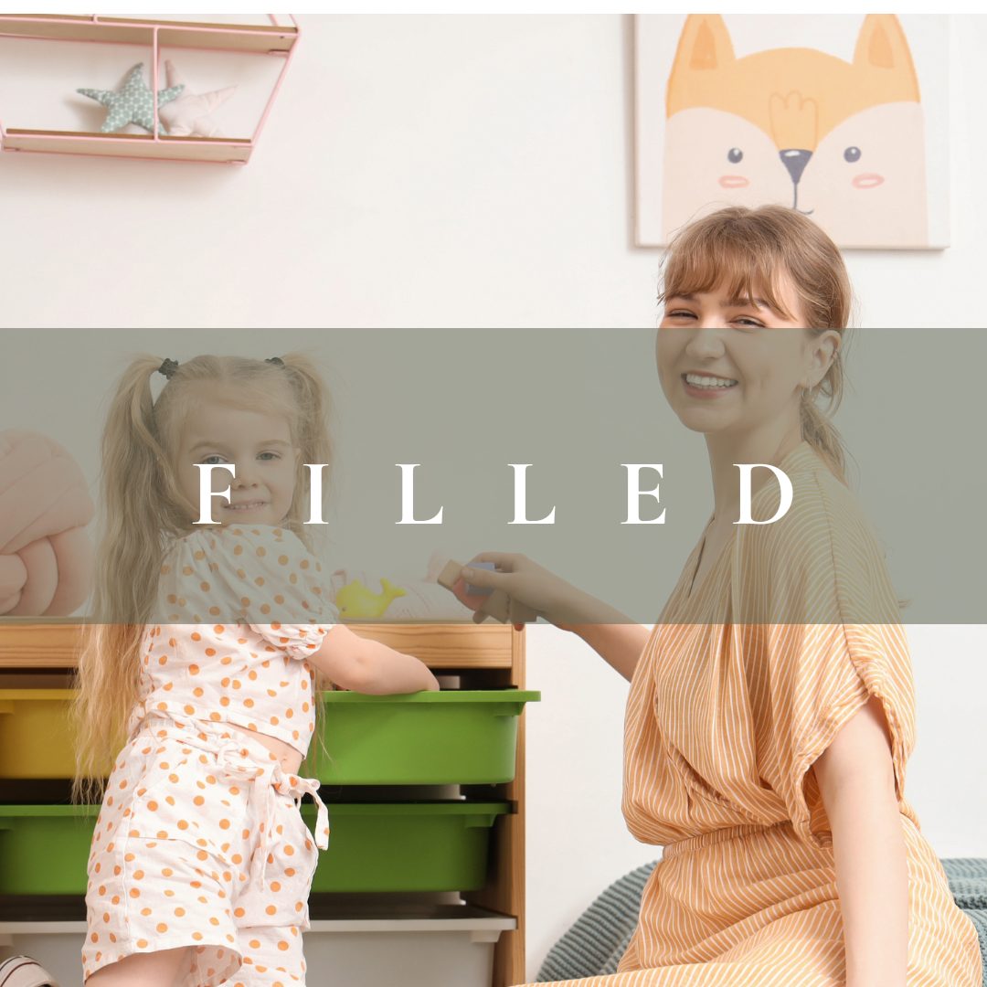 A young woman and a little girl playing together in a brightly lit room. The girl has blonde hair tied in pigtails and is wearing a white polka dot outfit. The woman is smiling and holding a toy, sitting on a couch. There are decorative items like a plush pillow and wall art with a fox face in the background.