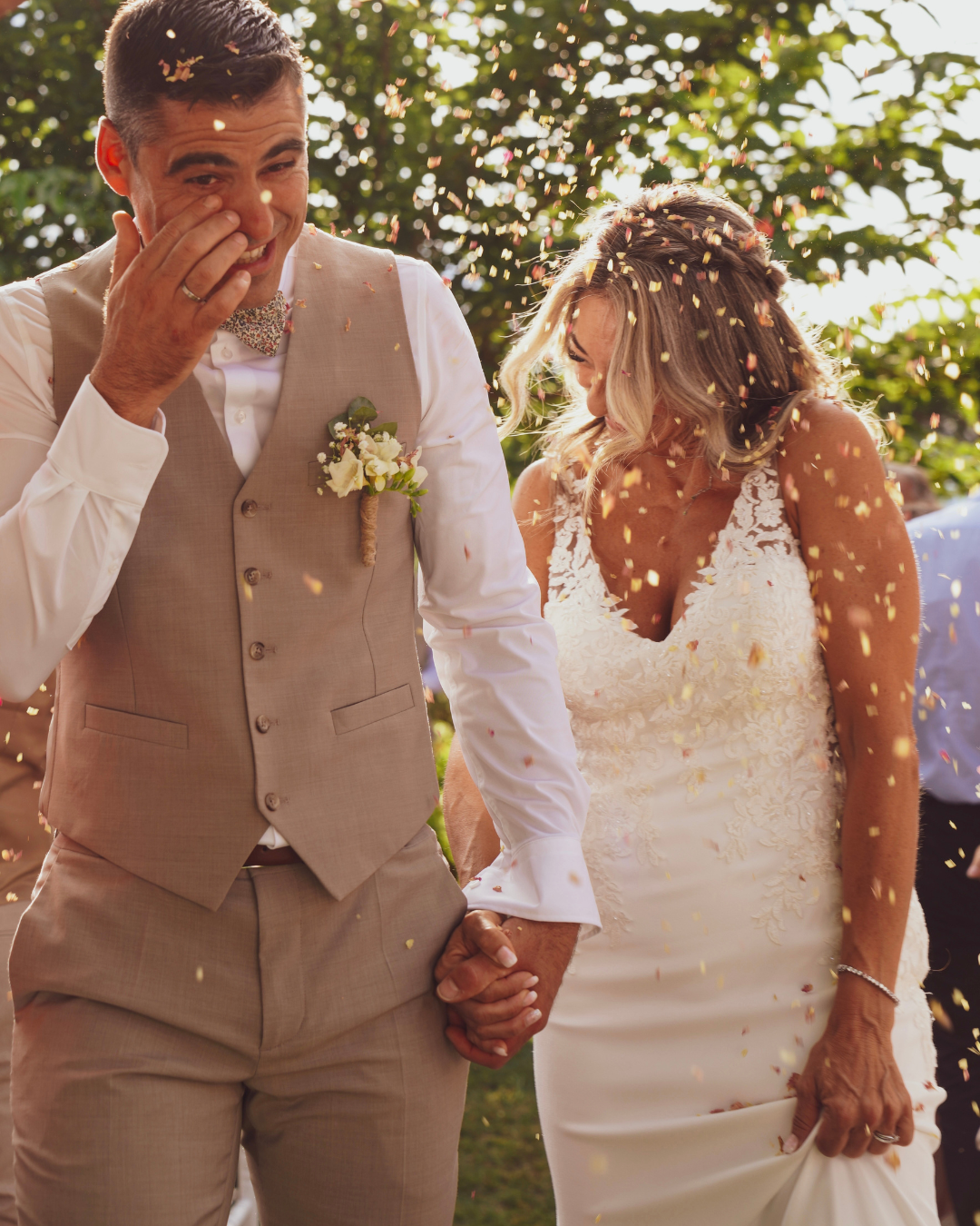 Newly married couple holding hands and smiling as confetti falls around them during an outdoor wedding celebration.