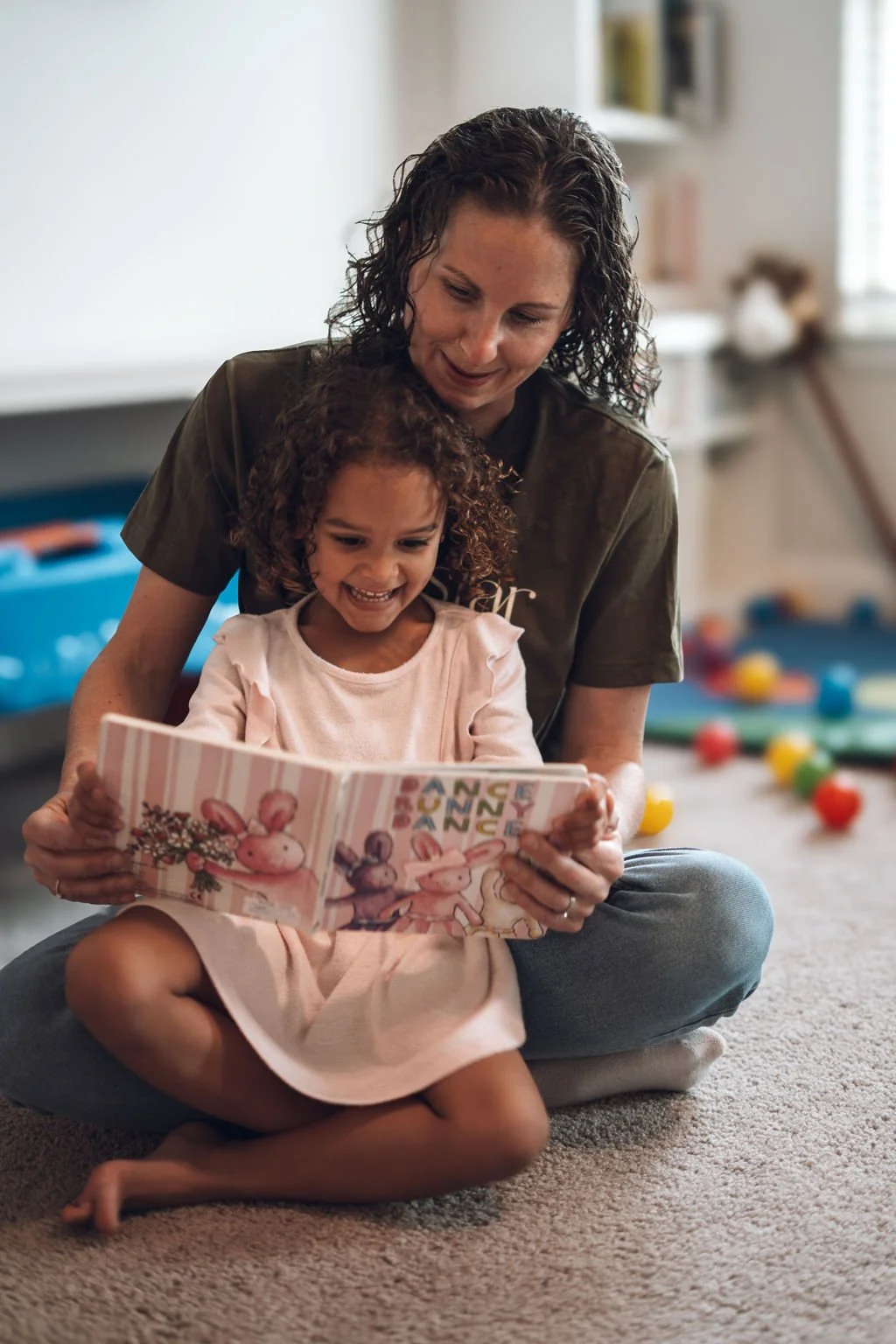 Woman sitting on the floor reading a children’s book with a young girl, both smiling and engaged in a cozy indoor playroom setting.