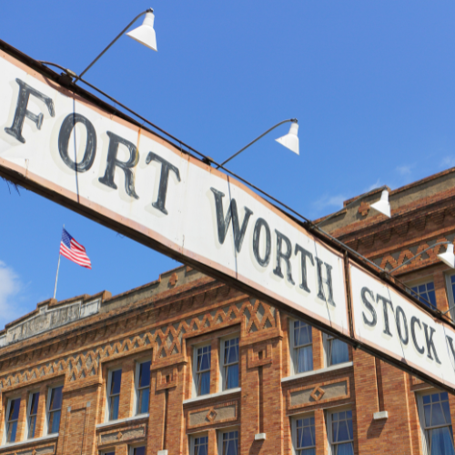 Fort Worth Stockyards sign mounted above a historic brick building with an American flag in the background under a clear blue sky.