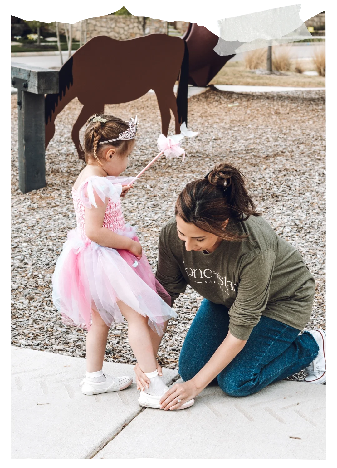 Woman kneeling outdoors helping a young girl in a pink dress and tiara adjust her shoe, with playground elements in the background.