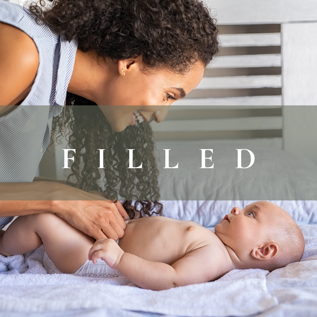 A smiling woman interacts with a baby lying on a bed, looking at each other. The woman is leaning over the baby, who is lying on their back on a white blanket in a bedroom setting.