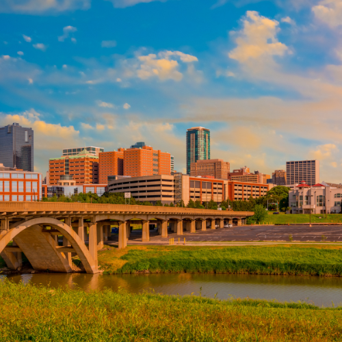 City skyline with a bridge crossing a river in the foreground, featuring modern buildings under a partly cloudy sky.