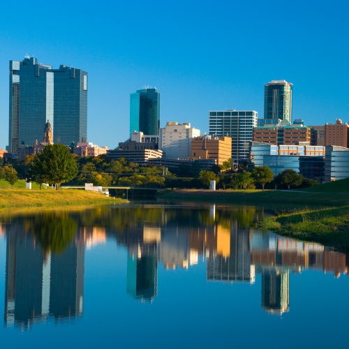 City skyline reflected in a calm body of water, with modern high-rise buildings and green parkland under a clear blue sky.