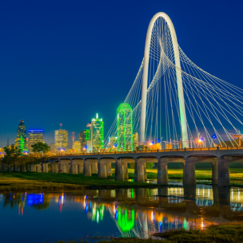 Margaret Hunt Hill Bridge illuminated at night with the Dallas skyline reflected in the Trinity River below.