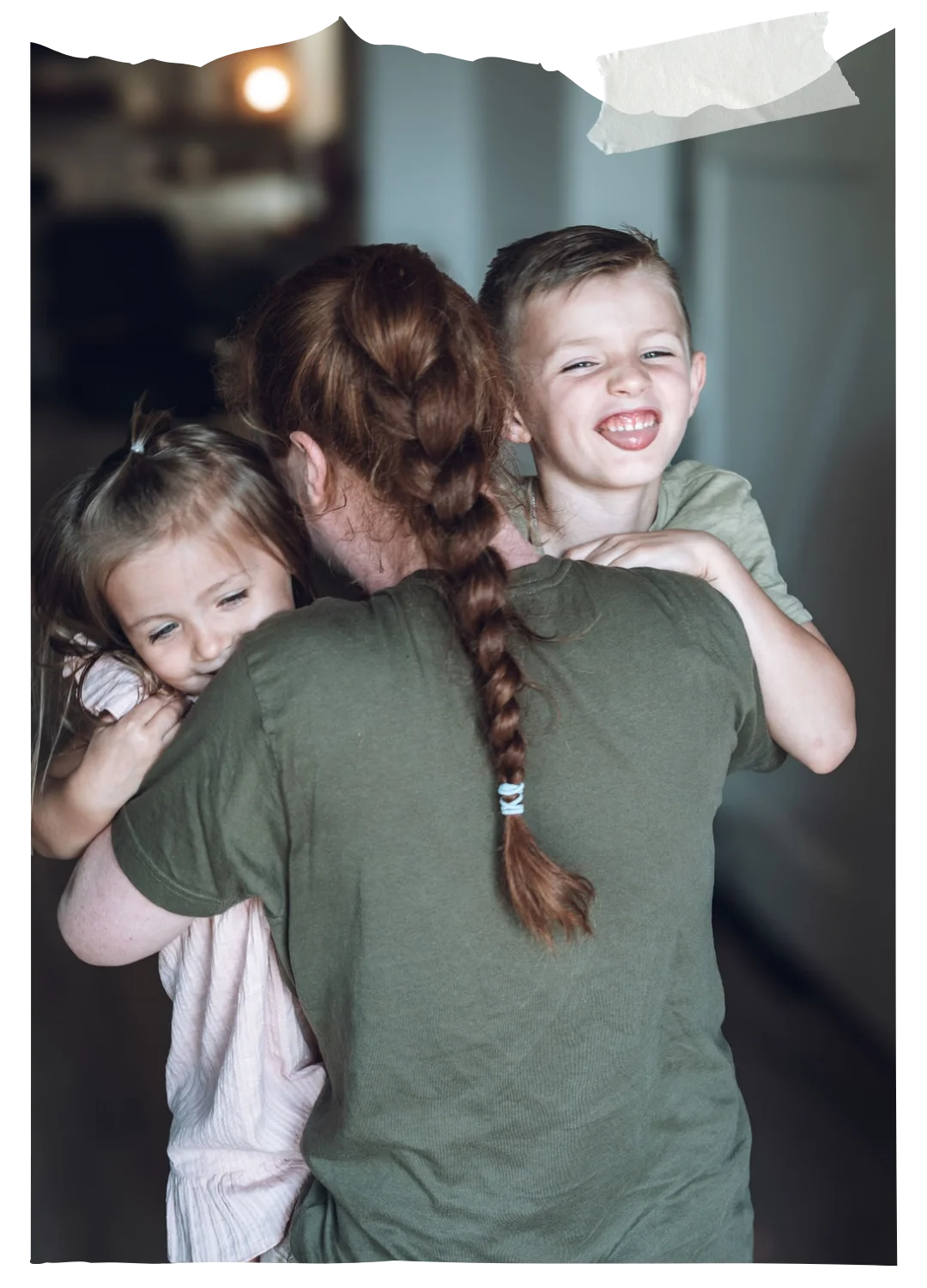 Woman with braided hair hugging two young children indoors, both smiling and showing affection in a warm family moment.
