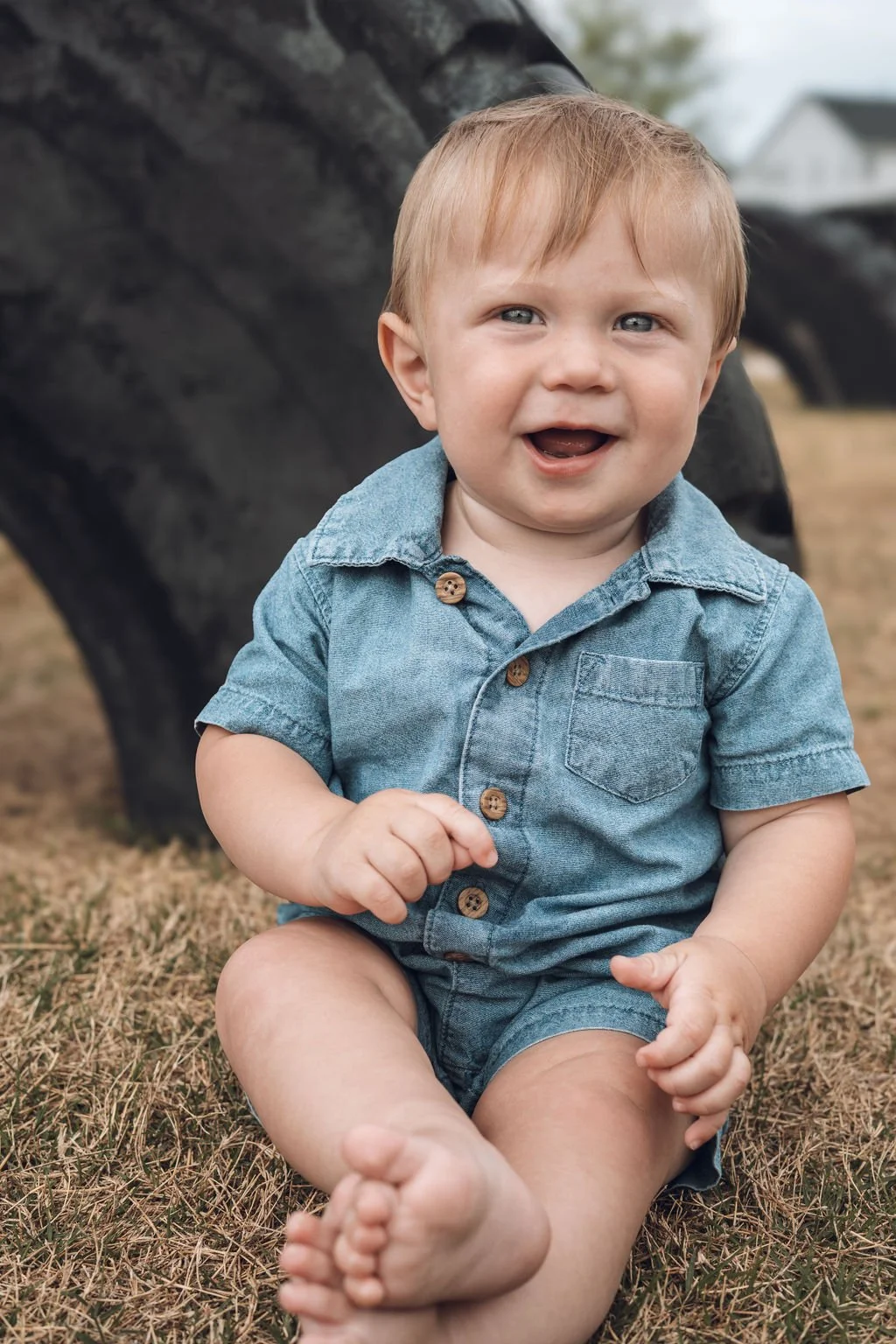 Smiling baby sitting on grass outdoors, wearing a blue outfit, with a blurred background and playful expression.
