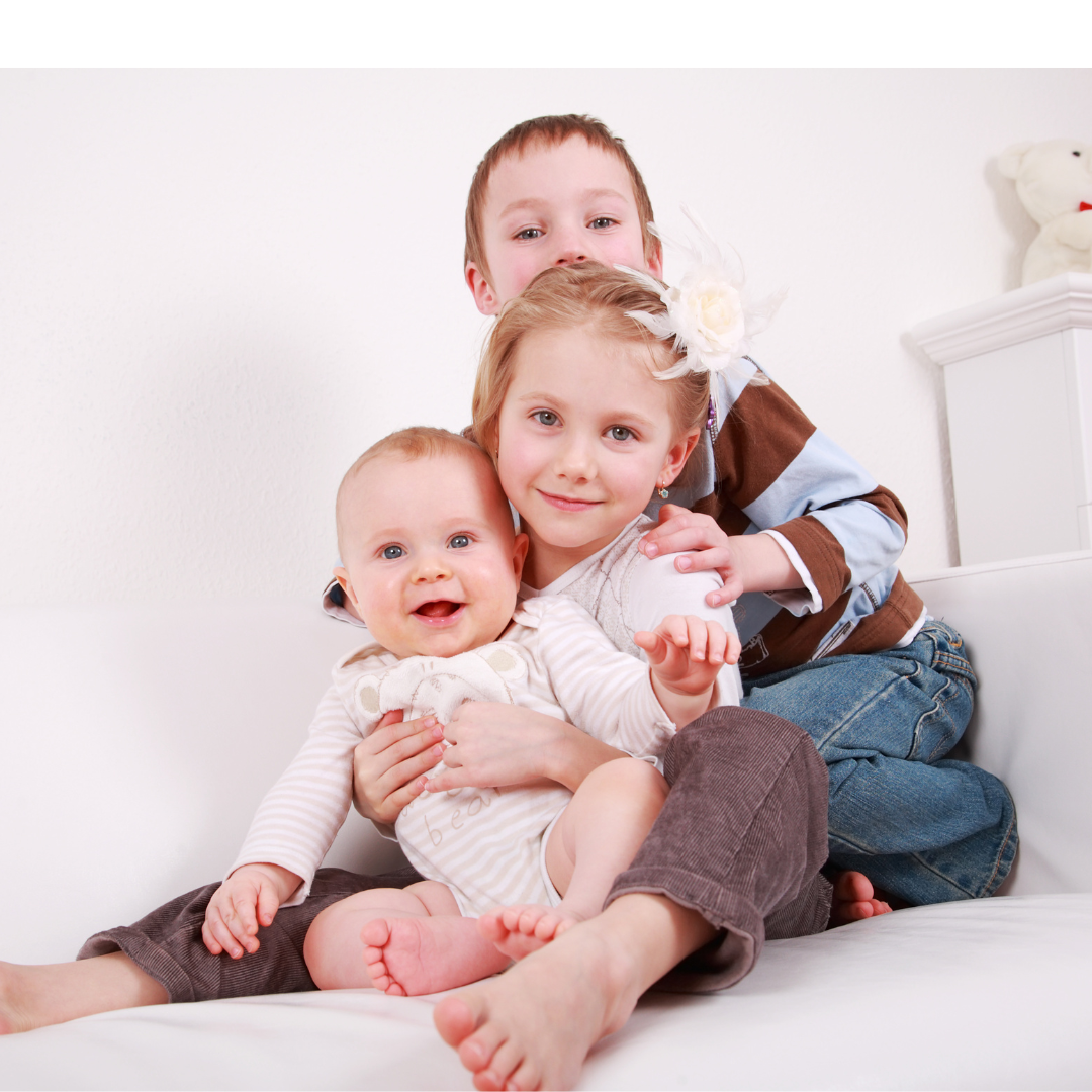 Three children sitting on a white couch, smiling at the camera with a white wall in the background.