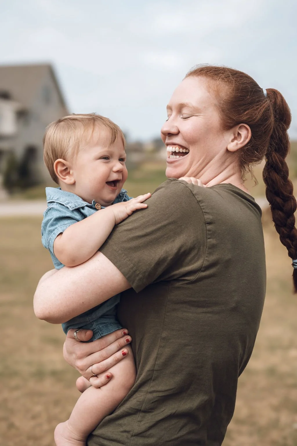 Woman holding a baby outdoors, both laughing and sharing a joyful moment with a blurred residential background.