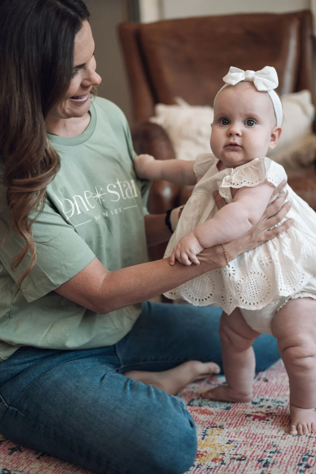 Woman sitting indoors helping a baby stand while holding their hands, both engaged in a warm and caring moment.