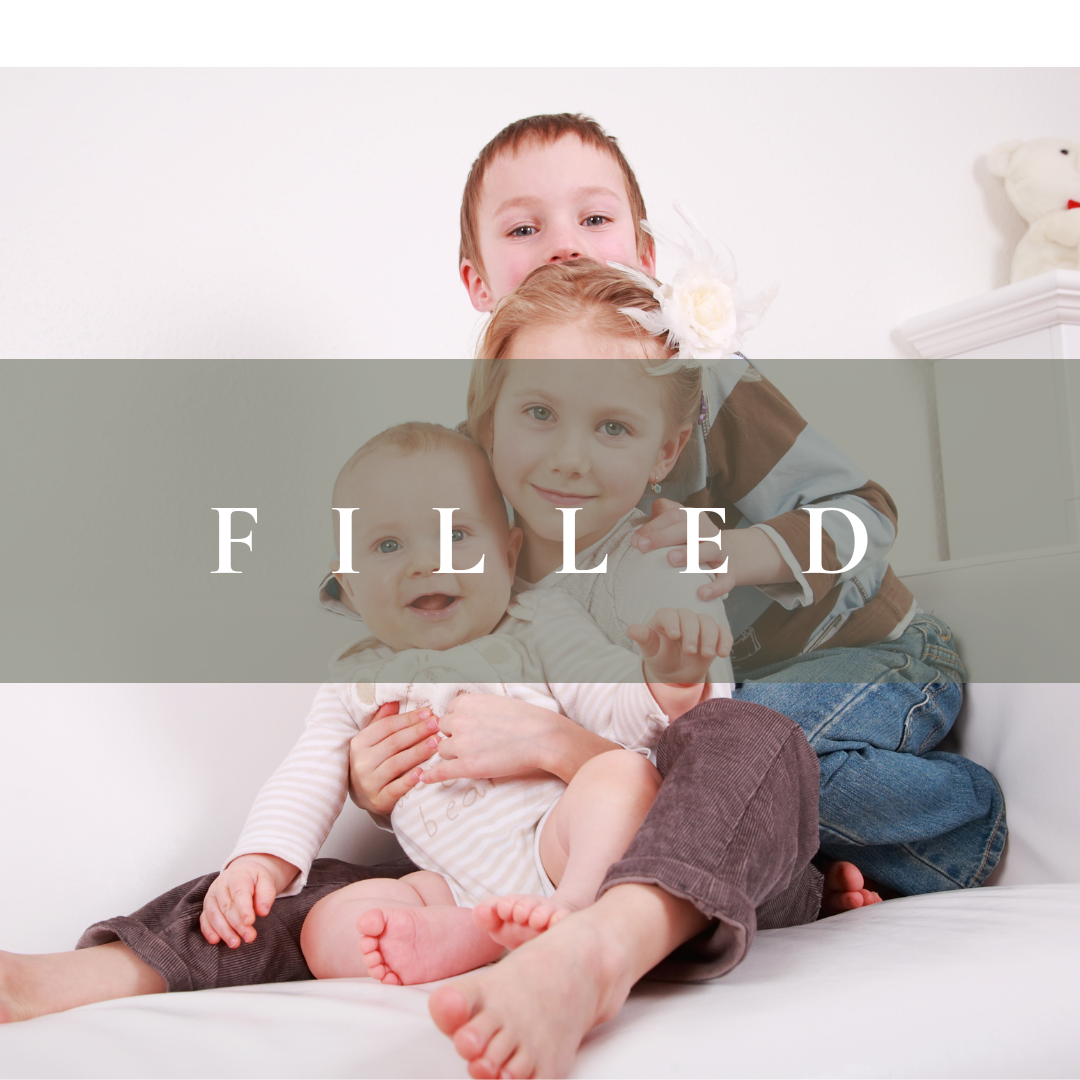 Three children sitting on a white couch, smiling at the camera with a white wall in the background.