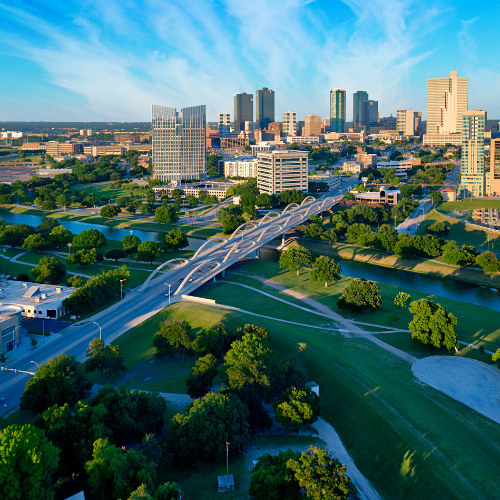 Aerial view of a city skyline with a modern bridge spanning a river, surrounded by green parks and buildings under a clear blue sky.