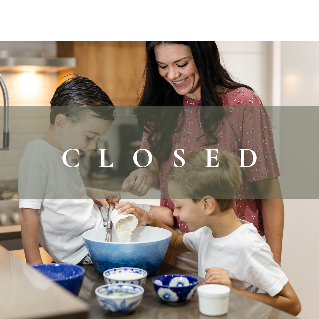 A woman and two young boys baking together in a kitchen, with one boy pouring flour into a blue mixing bowl while the other watches, all smiling and engaged in the activity.