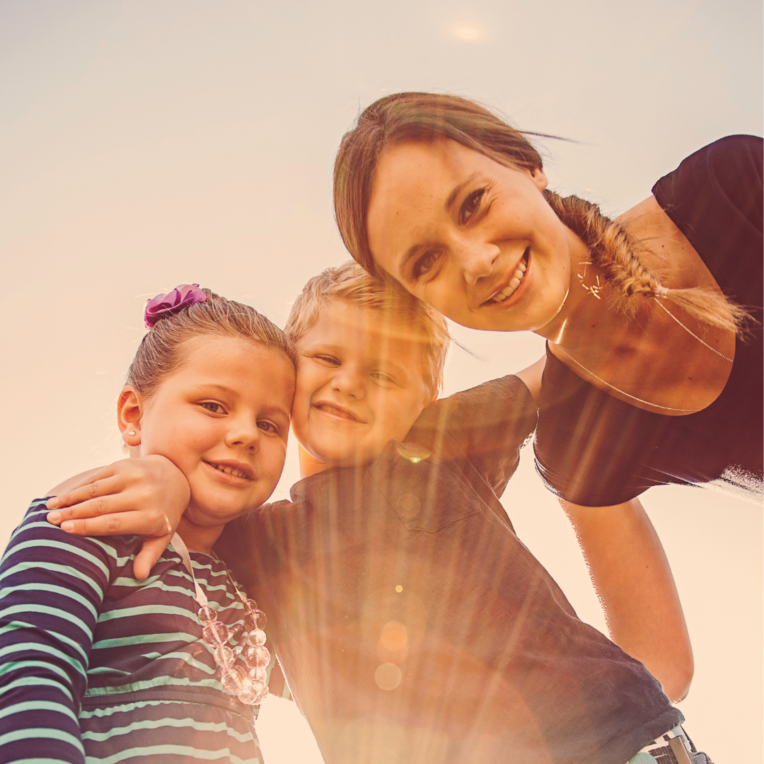 A woman and two children smiling and taking a selfie outdoors with the sun shining.