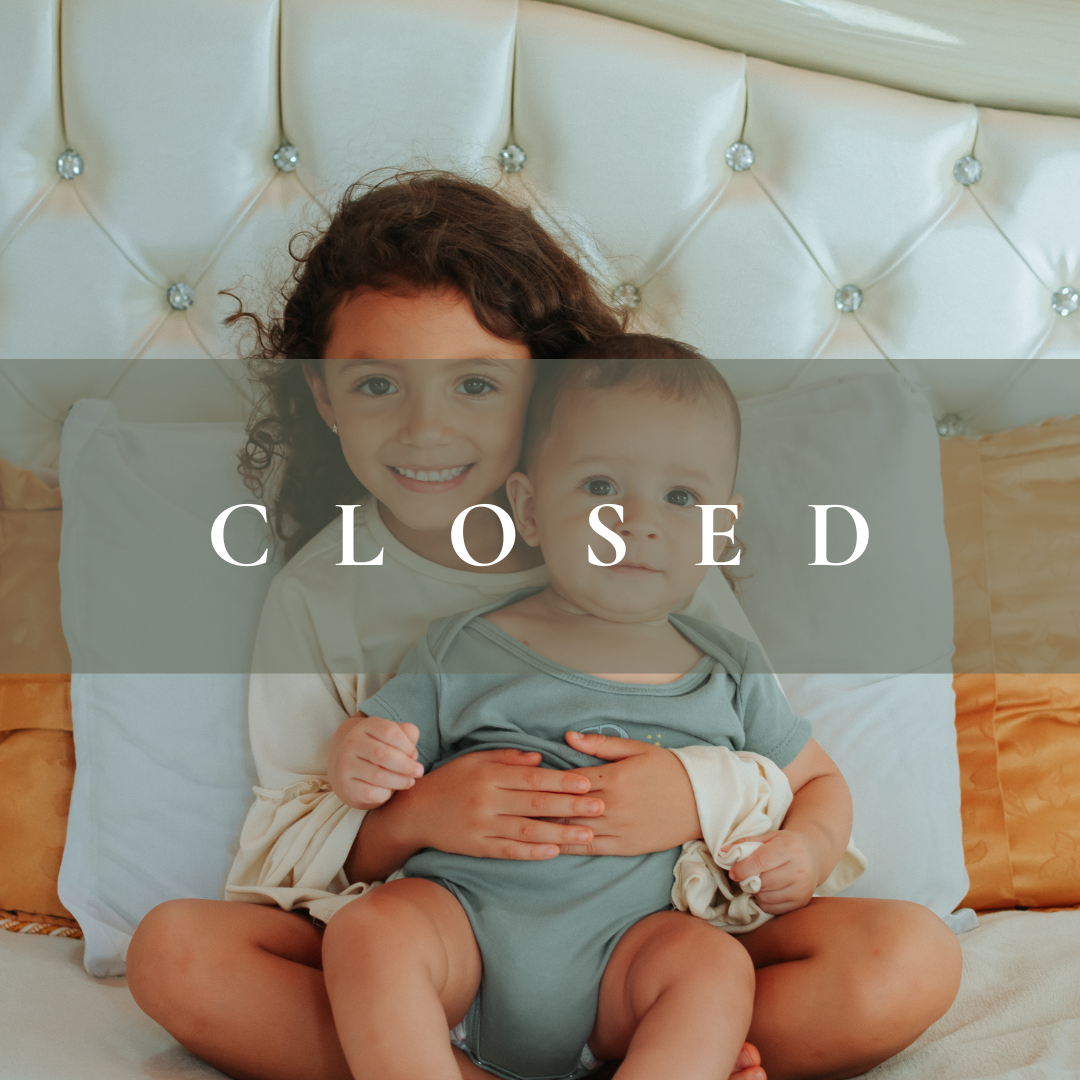 A young girl with curly hair sitting on a bed, holding a baby boy, both smiling at the camera, with a tufted white headboard and orange pillows behind them.