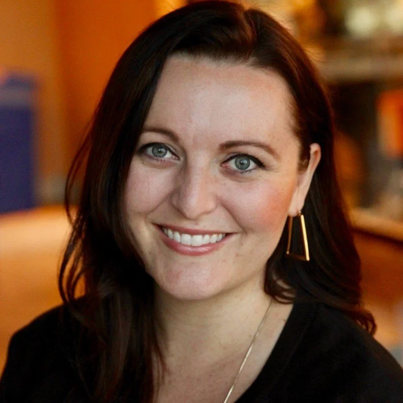 Close-up of a woman with long, dark brown hair, blue eyes, and a warm smile, wearing a black top and gold geometric earrings, in an indoor setting with warm lighting.