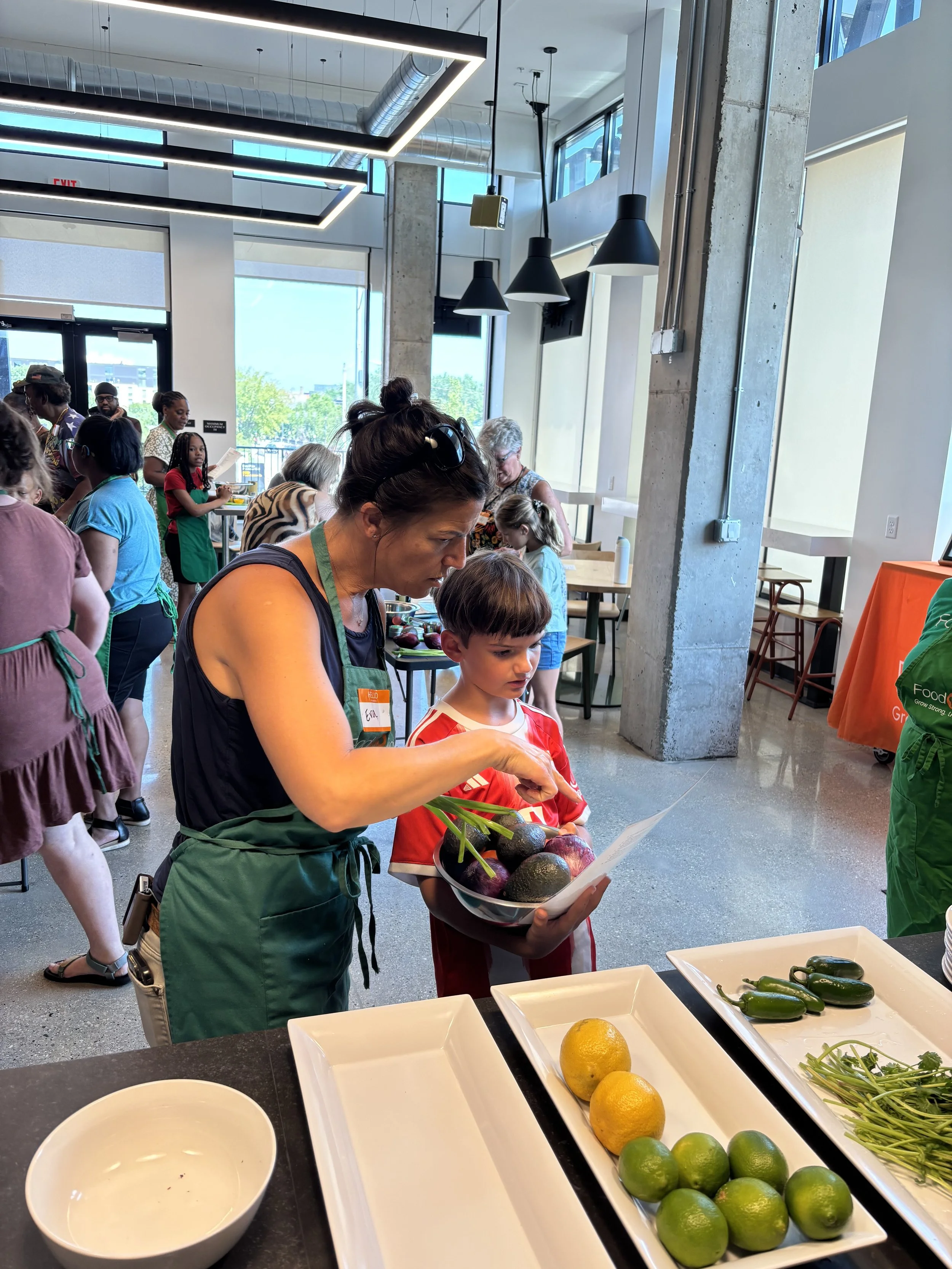People at a food event selecting vegetables and fruits at a market-style table in a modern building with large windows and contemporary lighting.