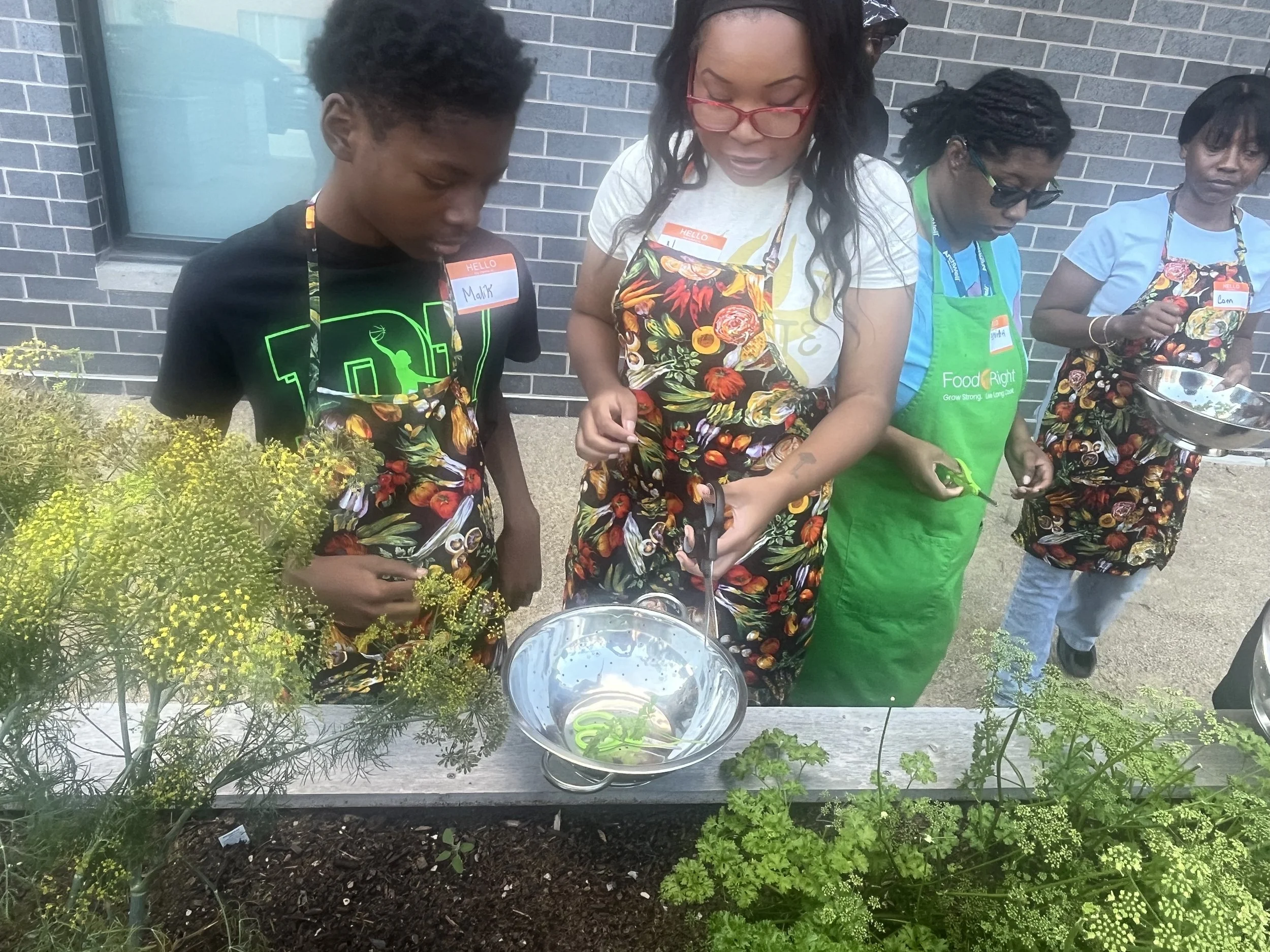 Four women wearing aprons and name tags stand behind a garden table with herbs and plants, preparing food outdoors against a brick wall.
