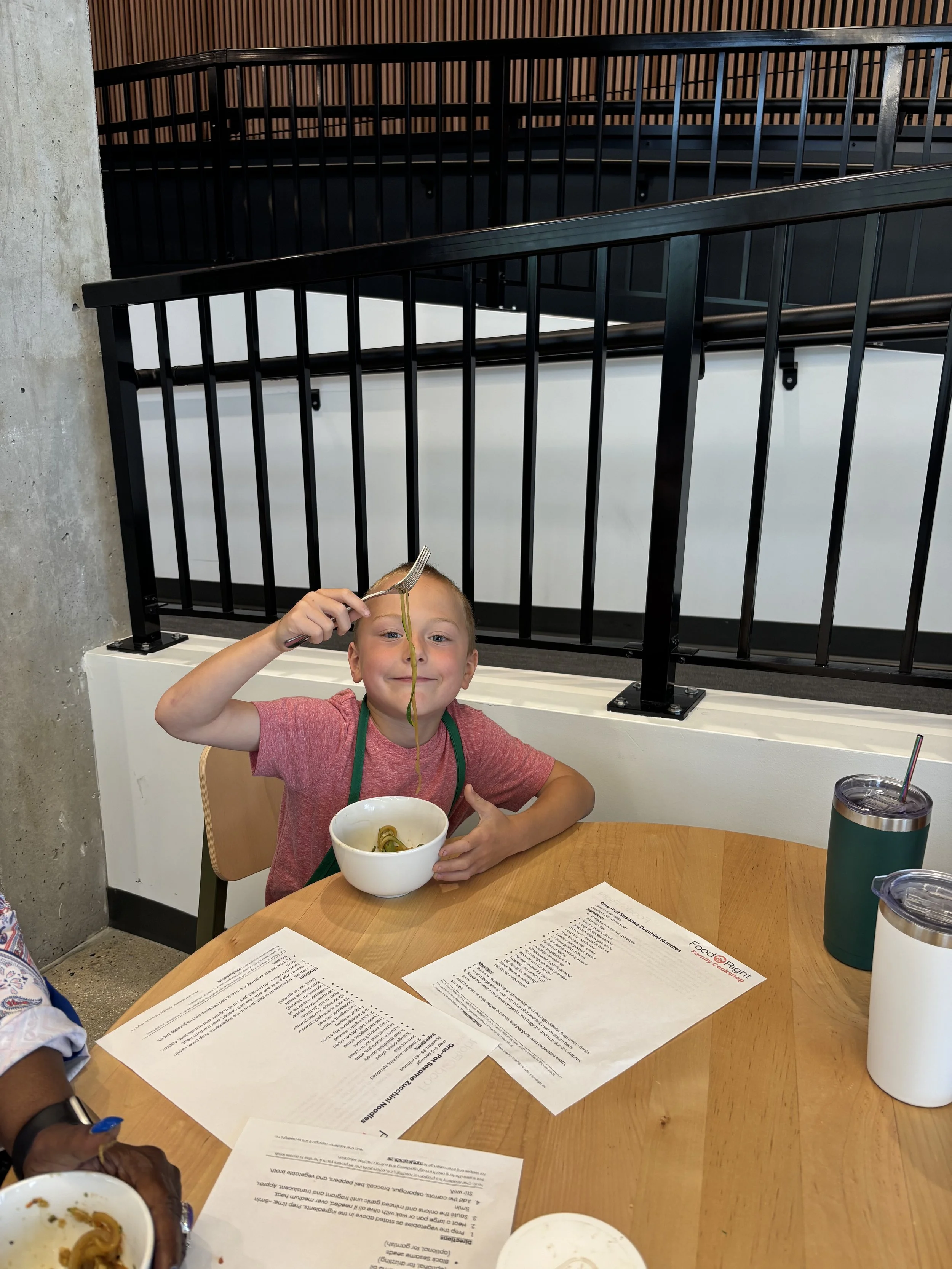 A young boy sitting at a round wooden table, holding a fork with noodles, with various papers and drinks on the table. The background features a staircase with black railings and a wooden wall.