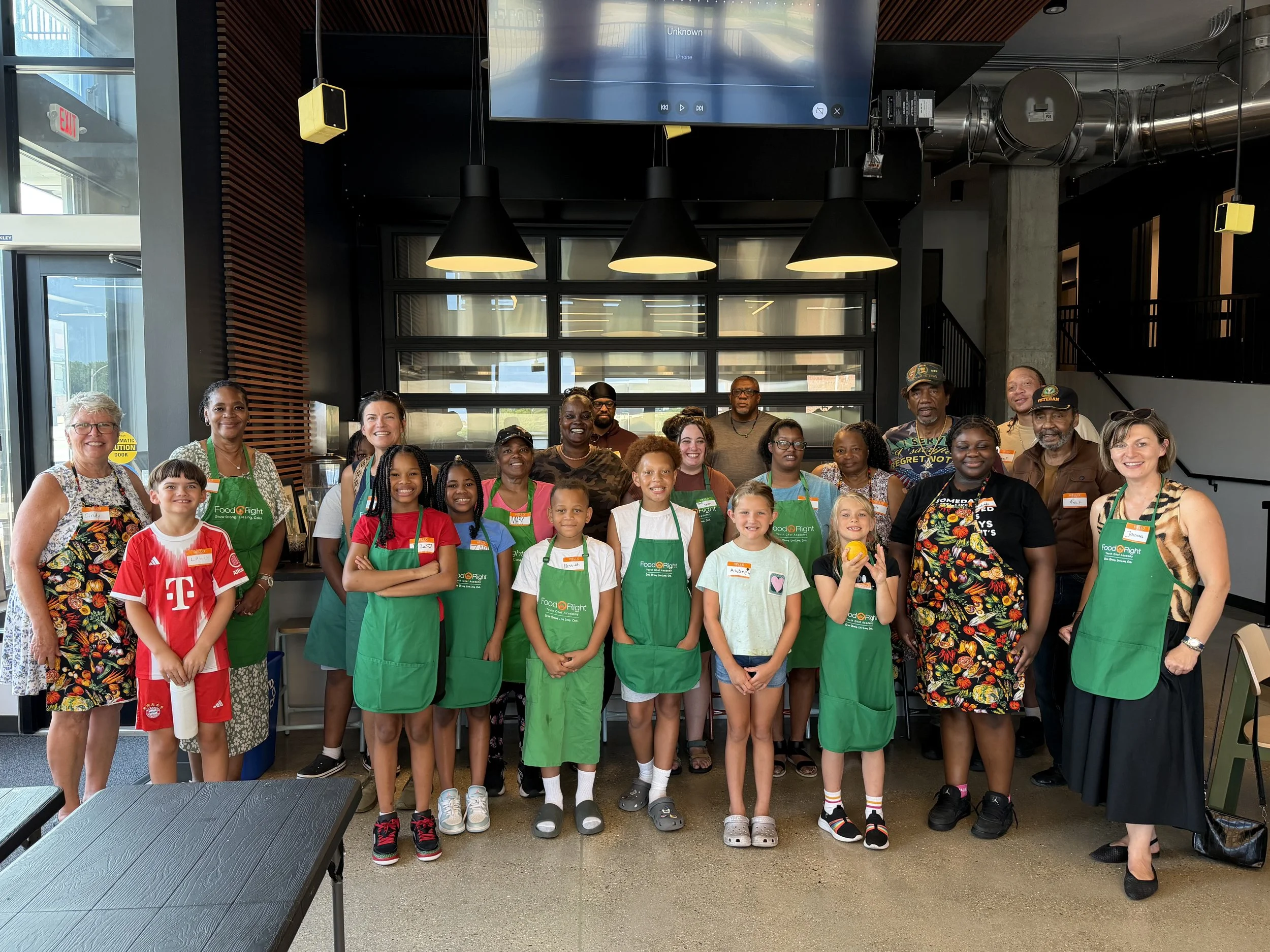 A group of diverse children and adults, some wearing green aprons, standing together inside a modern restaurant or cafe. They are smiling and posing for the photo, with a large glass wall and digital screen in the background.