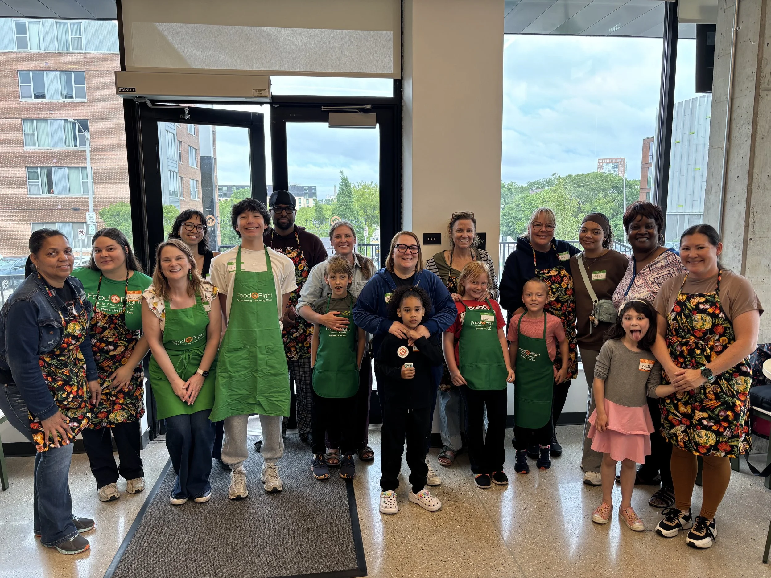 A diverse group of people, including children and adults, are standing together inside near the entrance of a building, smiling for a group photo. The background shows large windows with a view of neighboring buildings and trees outside.