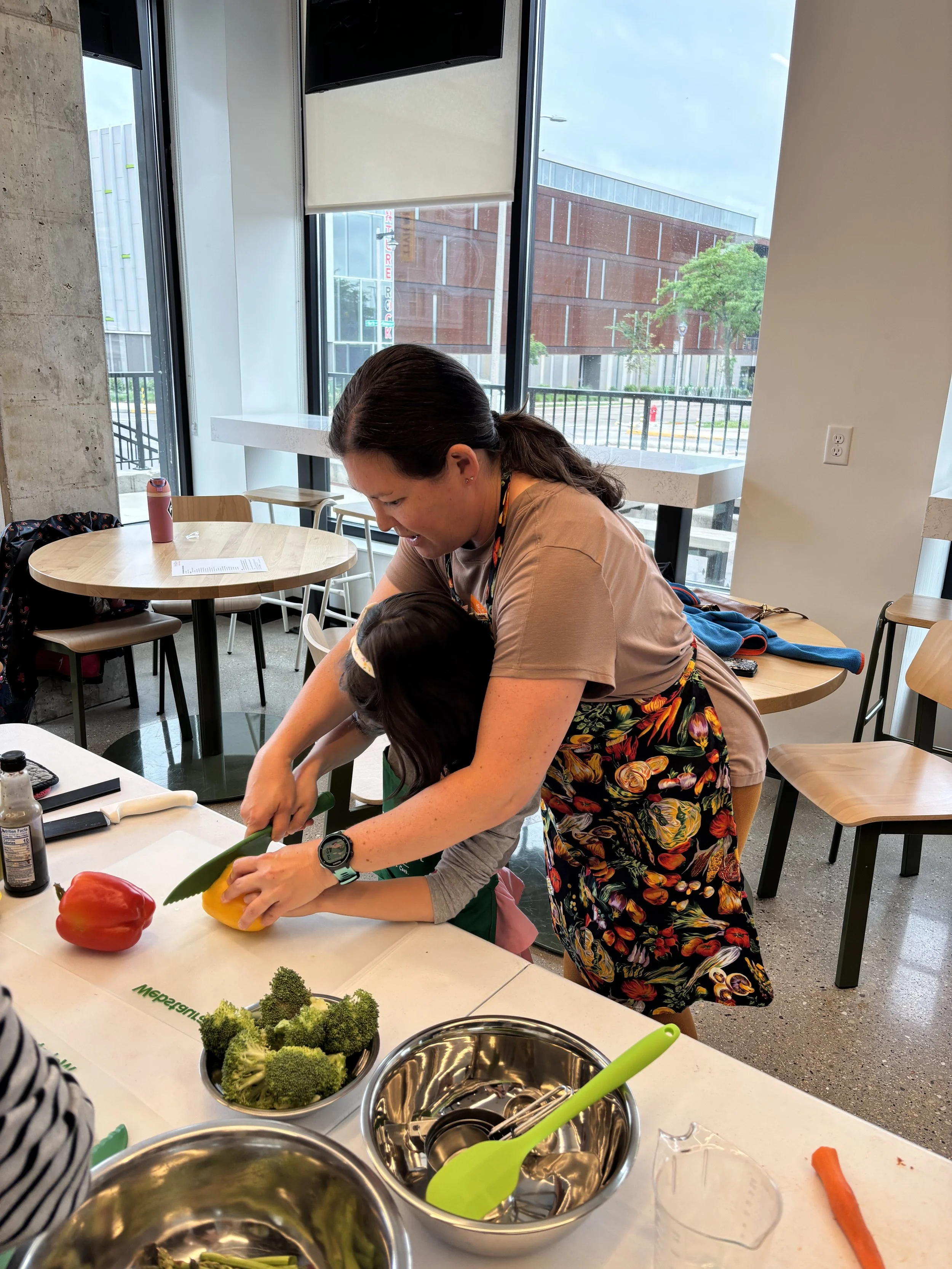 A woman assisting a child with preparing vegetables in a kitchen or cooking class setting, with various vegetables and kitchen tools on the table.